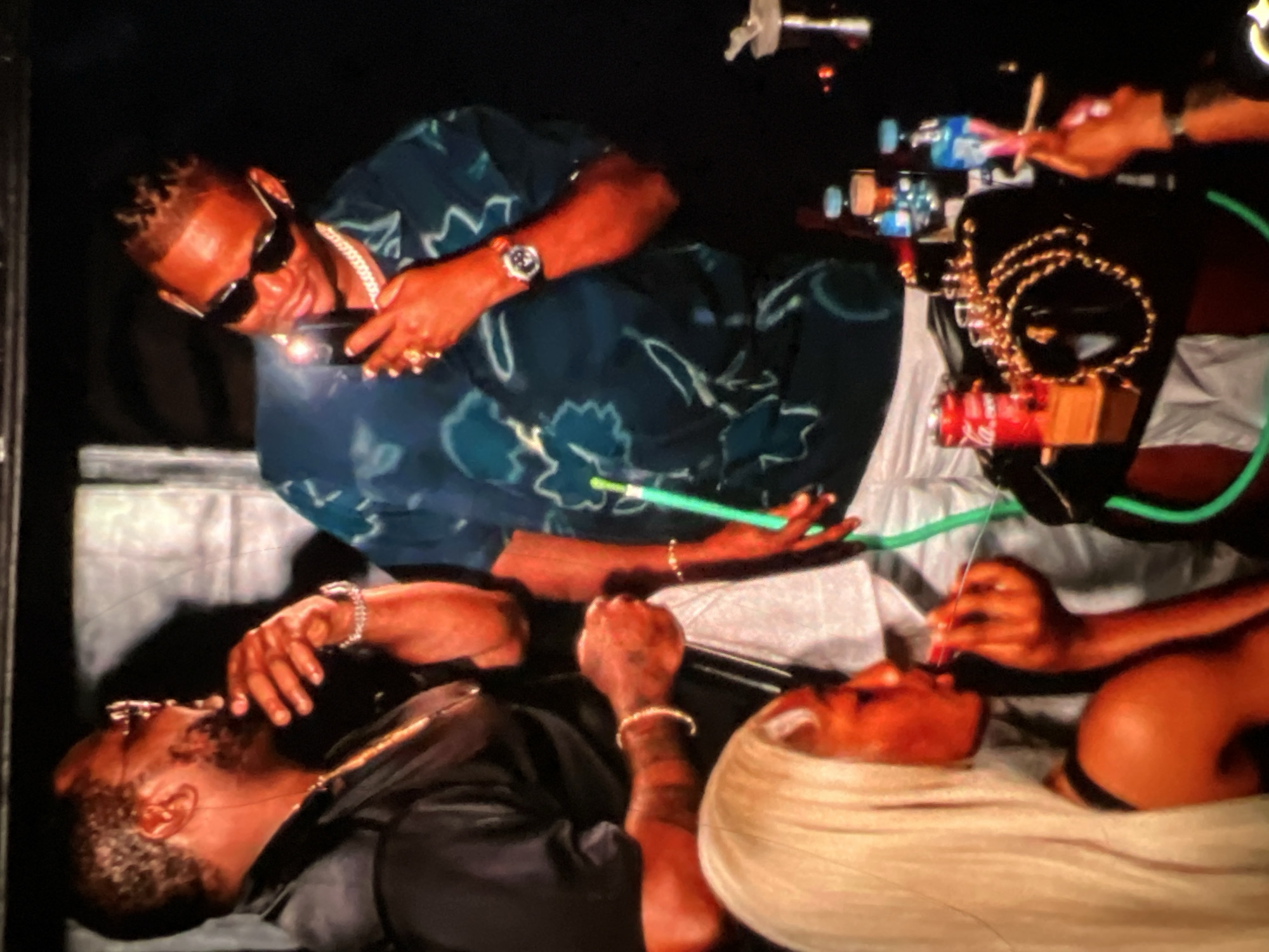 The image depicts an indoor scene, likely a club or a private party, with a dark, atmospheric setting. Three individuals are prominently visible, all appearing to be adults. On the left, a man with a beard and sunglasses is seen from the side, with his hand raised to his mouth, seemingly in conversation or contemplation. He wears a gold chain and a bracelet. To his right, another man, also wearing sunglasses and a patterned shirt, is holding a phone, possibly taking a picture or recording. He is adorned with rings and a watch. In the foreground, a woman with long blonde hair is partially visible, holding a red object to her lips, which resembles a lipstick or a similar cosmetic. The environment appears to be a social gathering, with various objects on a table in the background, including bottles of water, a can of Coca-Cola, and what looks like a hookah pipe with a green hose. The overall impression is one of leisure and social interaction. There is no visual information in the image to specifically place the scene within the city of Lagos, Nigeria.