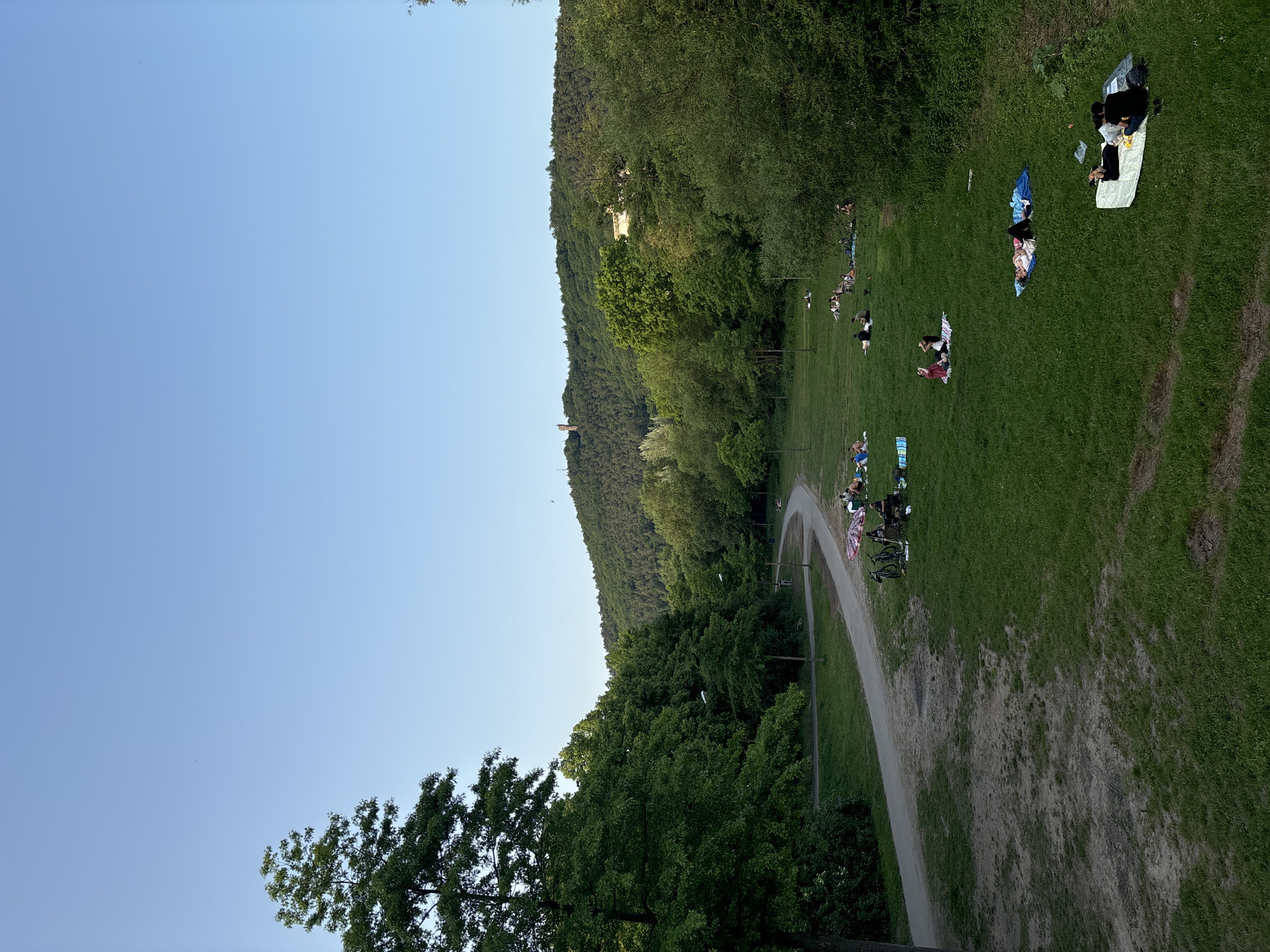 The image depicts a scene in Marburg, Germany, possibly along the Lahn River. A grassy area slopes upwards to a wooded hillside. A winding dirt path runs through the scene. A light-colored building, likely a historical tower or structure, is visible on the hillside. Multiple groups of people are scattered across the grass, relaxing and picnicking. Some are lying on blankets. Bicycles are parked near some groups. The sky is clear and blue. Trees and foliage are abundant, indicating a park or recreational area.