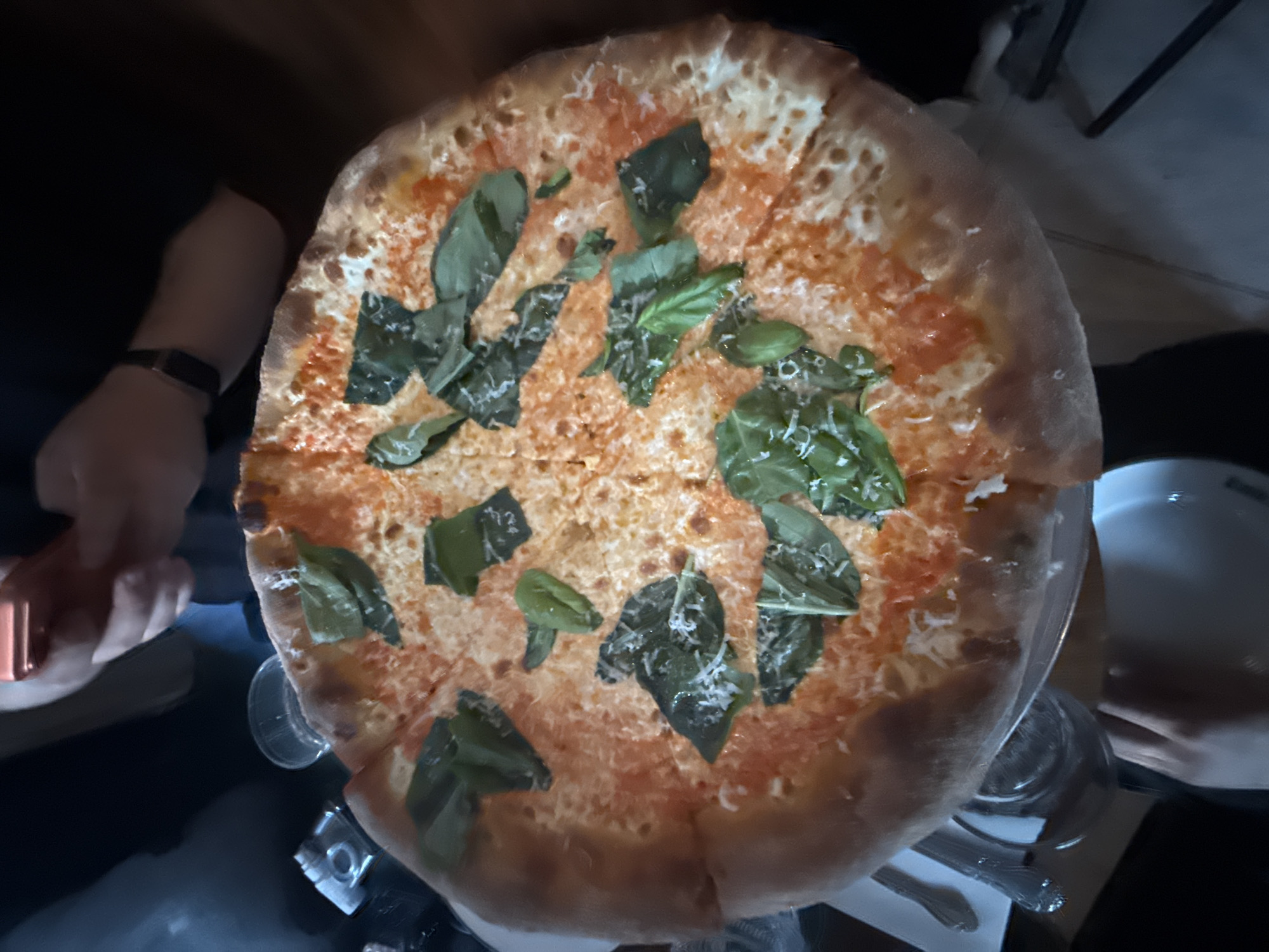 The image depicts a pizza, likely a Margherita variant, served on a clear stand. Fresh basil leaves are scattered across the pizza's surface. The pizza appears to be freshly baked, with a slightly charred crust. The image also captures the presence of individuals. A left hand with a watch is visible along with a right hand. Other objects include a small glass, a plate, and cutlery, hinting at a dining environment. The specific location is the city of New York, United States of America.