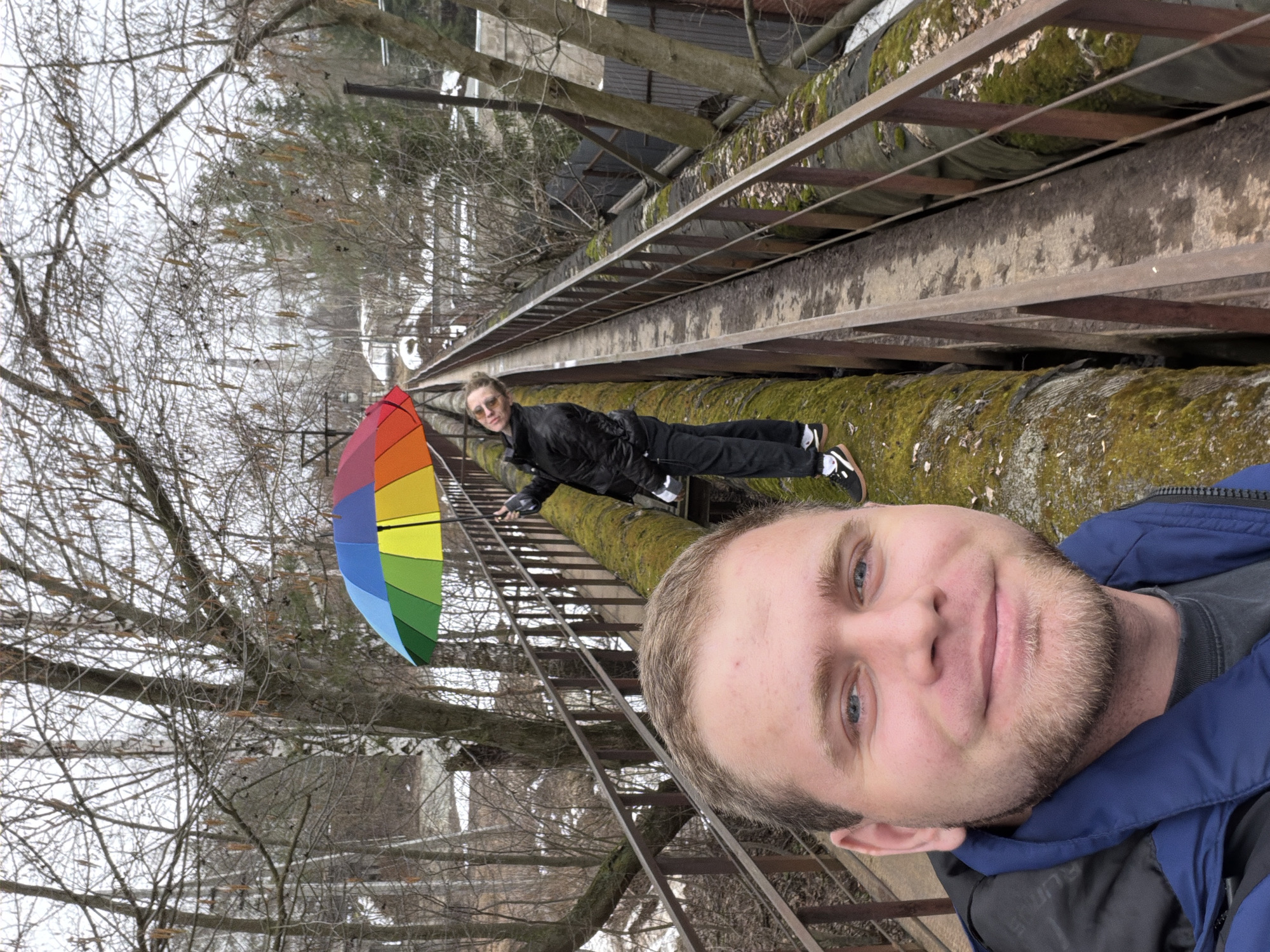The image is an eye-level selfie, taken by a man with blue eyes and light brown hair, wearing a dark gray shirt and a dark blue jacket. Another individual, wearing sunglasses and a black jacket and pants, is lying prone on a large, moss-covered pipe, stretching out. Between the two figures is a closed, multi-colored umbrella. The setting appears to be a large, above-ground pipe system, crisscrossed with metal support structures and ladders. Bare trees are visible, suggesting late autumn or winter, and what appears to be buildings are in the background. The location is Chaikovsky, Russia.