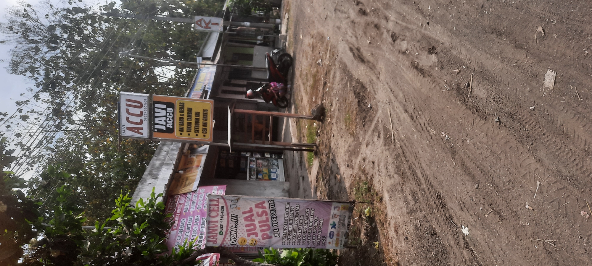 The image depicts a street scene in the village of Ngembul, Indonesia, where two prominent signs for "JAVI ACCU" are visible. The first sign, a vertical rectangular banner on the left, announces "JANVI CELL" and lists various services including "JUAL PULSA ALL OPERATOR," "PAKET DATA INTERNET," "VOUCHER INTERNET," "PAKET TELPON & SMS," "TOKEN LISTRIK," "TOP UP DANA, OVO," "TOP UP SHOPEEPAY," "TOP UP GOPAY," and "TOP UP GAME." The second sign, a smaller square banner above "JAVI ACCU" at the top right, also displays "JAVI ACCU" with a phone number "082245301631". Below this, a larger yellow sign for "JAVI ACCU" lists services such as "AKI BARU & BEKAS" (New and Used Batteries), "TUKAR TAMBAH" (Trade-in), "SETROOM AKI" (Battery Charging), and "CEK AKI GRATIS" (Free Battery Check). A red motorcycle with a rider wearing a helmet is parked in front of the buildings. The environment appears to be a commercial area with several small shops, and the ground is a dirt road with tire tracks. Trees and power lines are visible in the background.