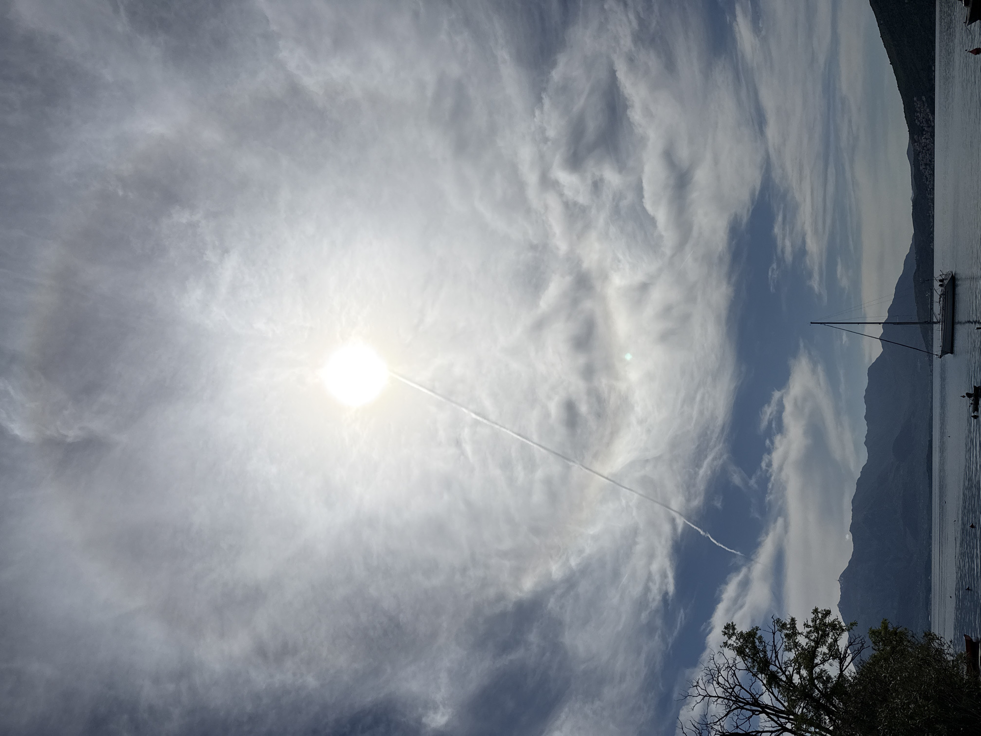The image, correctly oriented as a landscape, captures a serene outdoor scene over a body of water in Đenovići, Montenegro. The sky dominates the upper and left sections, filled with wispy, high-altitude cirrus clouds that create a soft, hazy diffusion of light.
The sun shines brightly from the upper-left, its intensity creating a luminous glow and a subtle solar halo around it. A long, white contrail from an aircraft cuts diagonally across the sky, passing near the sun.
On the right side, a calm body of water, likely a bay or a lake, reflects the soft light from the sky. Beyond the water, a series of dark, undulating mountains rise, their silhouettes creating a dramatic backdrop against the lighter sky.
A single-masted sailboat is visible on the water, anchored or moored near the base of the mountains, its mast reaching upwards. In the lower-right foreground, the dark, bare branches of a tree and some green foliage are visible, partially obscuring the view.
The bright sunlight suggests it is late morning or early afternoon. The weather appears calm with high clouds, and no specific activity or people are visible. There is no discernible text in the image.