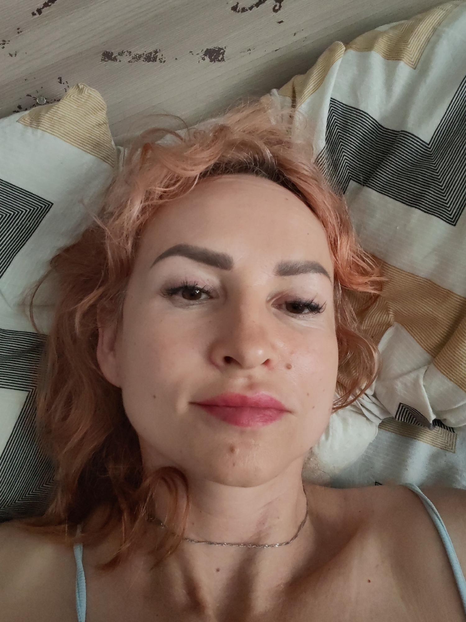 An overhead close-up shot shows a woman with light brown, wavy hair, lying on a bed with a patterned duvet. The woman is wearing light blue shoulder straps and a silver necklace. Her eyes are open, and she is wearing makeup, including pink lipstick and mascara. The background consists of a wooden surface and the patterned duvet. No specific location in Chaikovsky, Russia, is discernible.