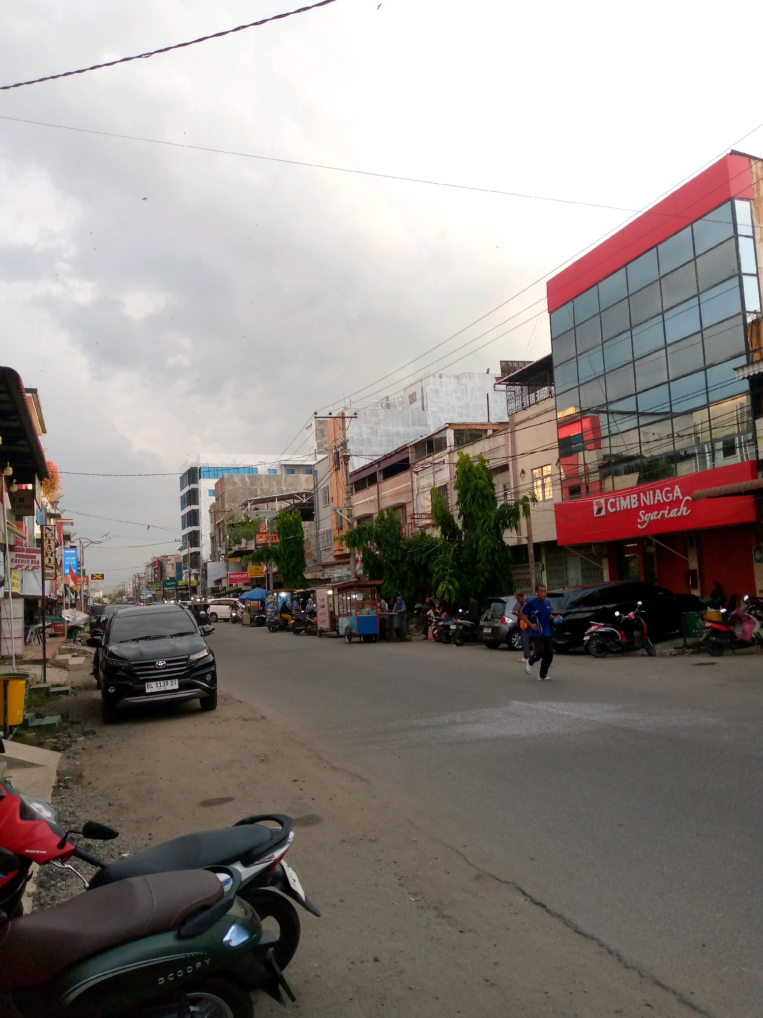 A street view in Lhokseumawe, Indonesia, shows a two-lane asphalt road bordered by buildings and parked vehicles. On the right side of the street, a modern building with a red facade and glass windows displays the "CIMB NIAGA Syariah" logo. Several cars and motorcycles are parked along the road. A lone individual is running on the road. On the left side of the street, there are more businesses, including a notary office, and parked motorcycles. The sky is overcast, and there are visible utility wires overhead. In the foreground, two parked scooters are visible.