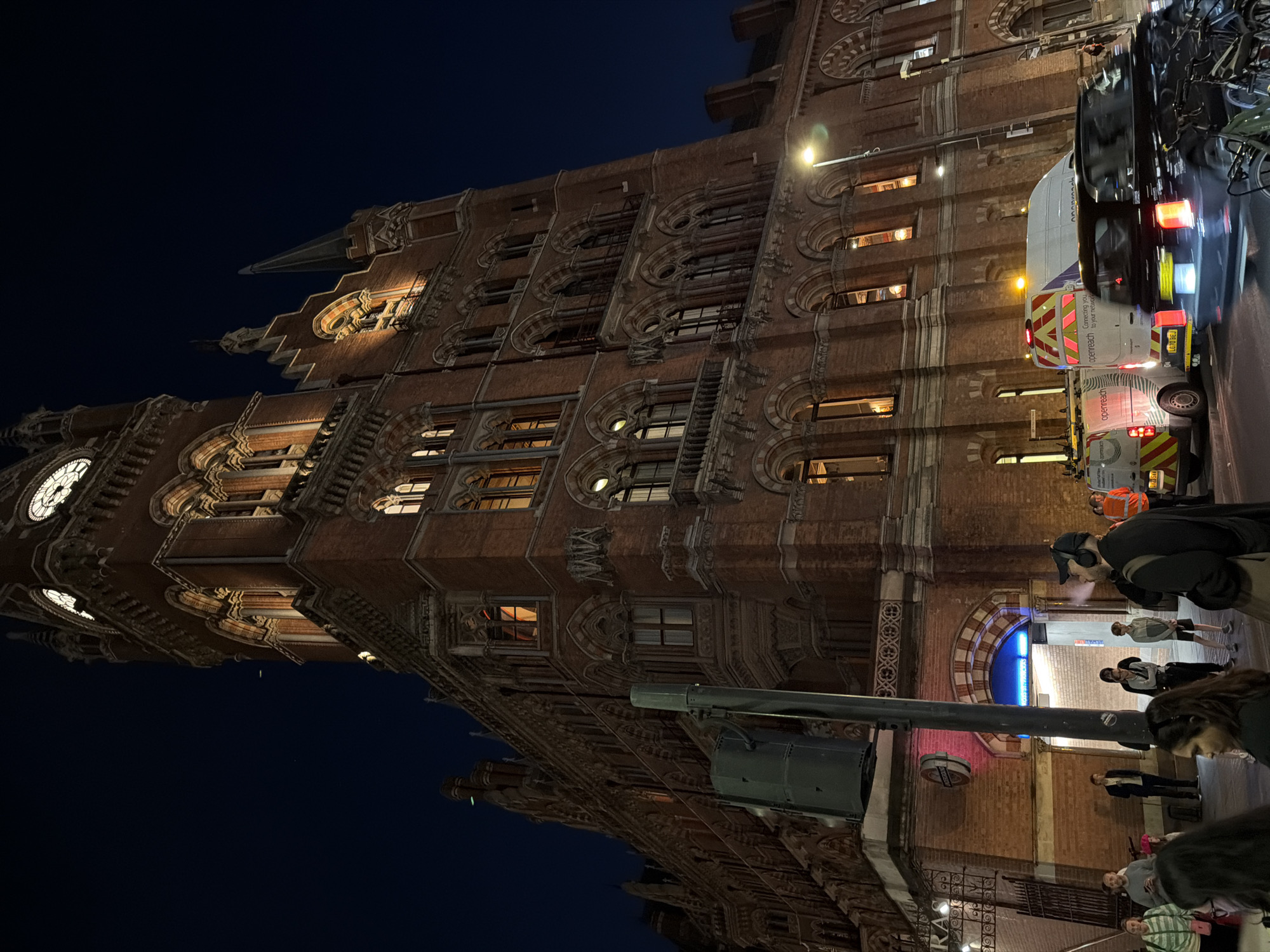 The image shows St. Pancras International train station in London at night. The building's facade is illuminated, highlighting its architectural details. People are visible on the sidewalk in front of the station entrance. Several utility vehicles can be seen parked on the street alongside the building. A street light and a traffic signal are also visible.