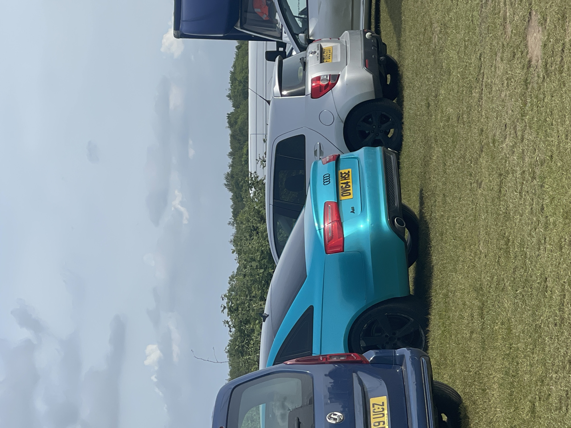The image captures a row of parked cars on a grassy field, likely in the city of Sevenoaks, United Kingdom. The cars include a blue Vauxhall, a teal Audi A3 with license plate OV64 HSE, a silver hatchback, and a white van. The background consists of a treeline against a partly cloudy sky. The scene appears to be outdoors during daylight hours.