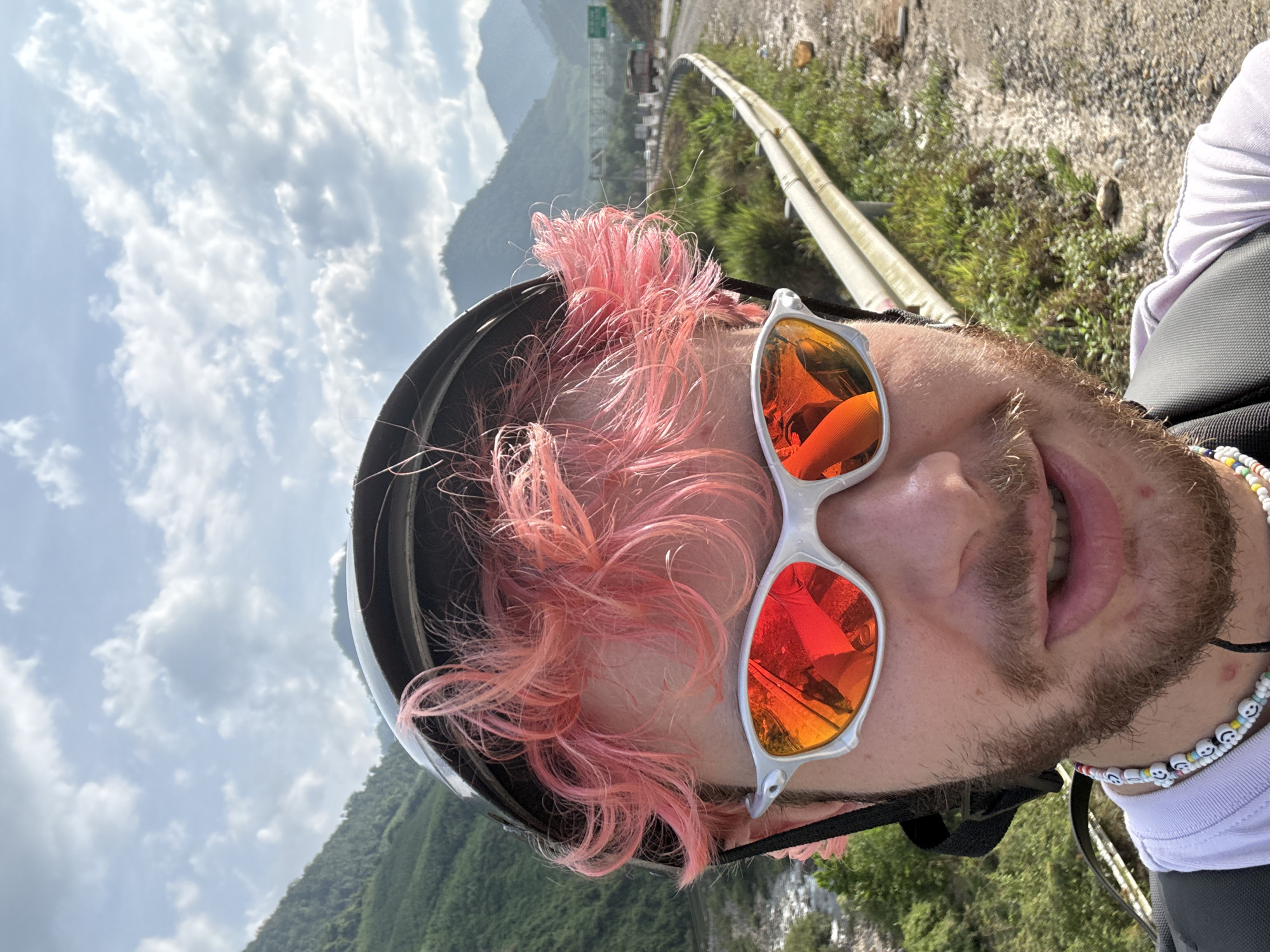 A close-up, slightly rotated selfie shows a man wearing a black helmet, white-framed sunglasses with orange reflective lenses, and a white shirt. His hair is dyed pink. He is wearing a colorful beaded necklace. He has a beard and mustache.
In the background, a mountain range is visible under a cloudy sky. A highway with a metal guardrail runs alongside a hillside covered in green vegetation. The location is possibly in the city of Đà Nẵng, Vietnam, inferred from the presence of a mountain range and lush vegetation, although this cannot be definitively confirmed from the image alone.