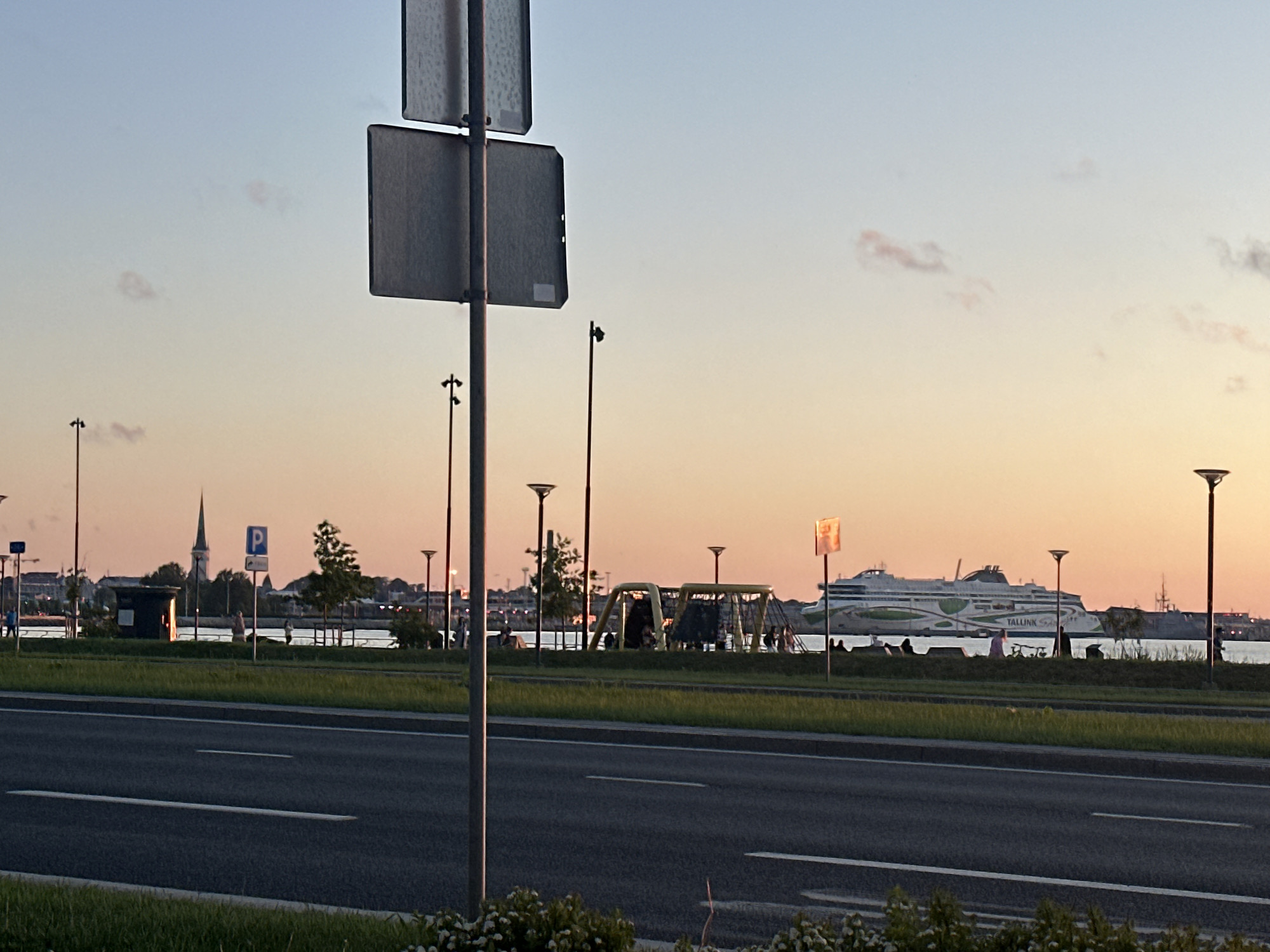 This image depicts a coastal scene in Tallinn, Estonia, likely at sunset or sunrise. The sky exhibits a gradient from light blue at the zenith to soft orange and pink hues near the horizon, with a few scattered, wispy clouds.
In the foreground, a multi-lane asphalt road with distinct white lane markings runs horizontally across the bottom third of the frame. A grassy median strip, featuring low-lying green vegetation and some white flowering plants, separates the road from the waterfront. A prominent grey metal signpost, holding two blank rectangular signs, stands upright on the median strip in the left-center of the image.
The midground features a waterfront promenade. From left to right:
* A small, dark building or kiosk, illuminated from within, stands on the promenade.
* Further back, a tall, dark church spire with a pointed roof is visible against the skyline, indicative of Tallinn's Old Town architecture (e.g., St. Olav's Church).
* A blue "P" (parking) sign on a pole is situated near the building.
* Numerous tall streetlights with illuminated tops line the promenade.
* Several indistinct human figures are visible along the promenade, some walking, others standing or possibly sitting on benches.
* A yellow-framed playground structure with climbing nets is present in the central section of the promenade.
* A large white ferry, clearly branded "TALLINK" with green decorative elements, is positioned on the water in the right midground. Partial lettering, possibly "SARA" or "STAR", is discernible on its hull.
* Other darker, more distant ships or port structures are visible beyond the Tallink ferry.
The background consists of the distant silhouette of Tallinn's cityscape and other industrial port elements against the colorful horizon. The calm water of the harbor separates the promenade from the ships and distant structures.