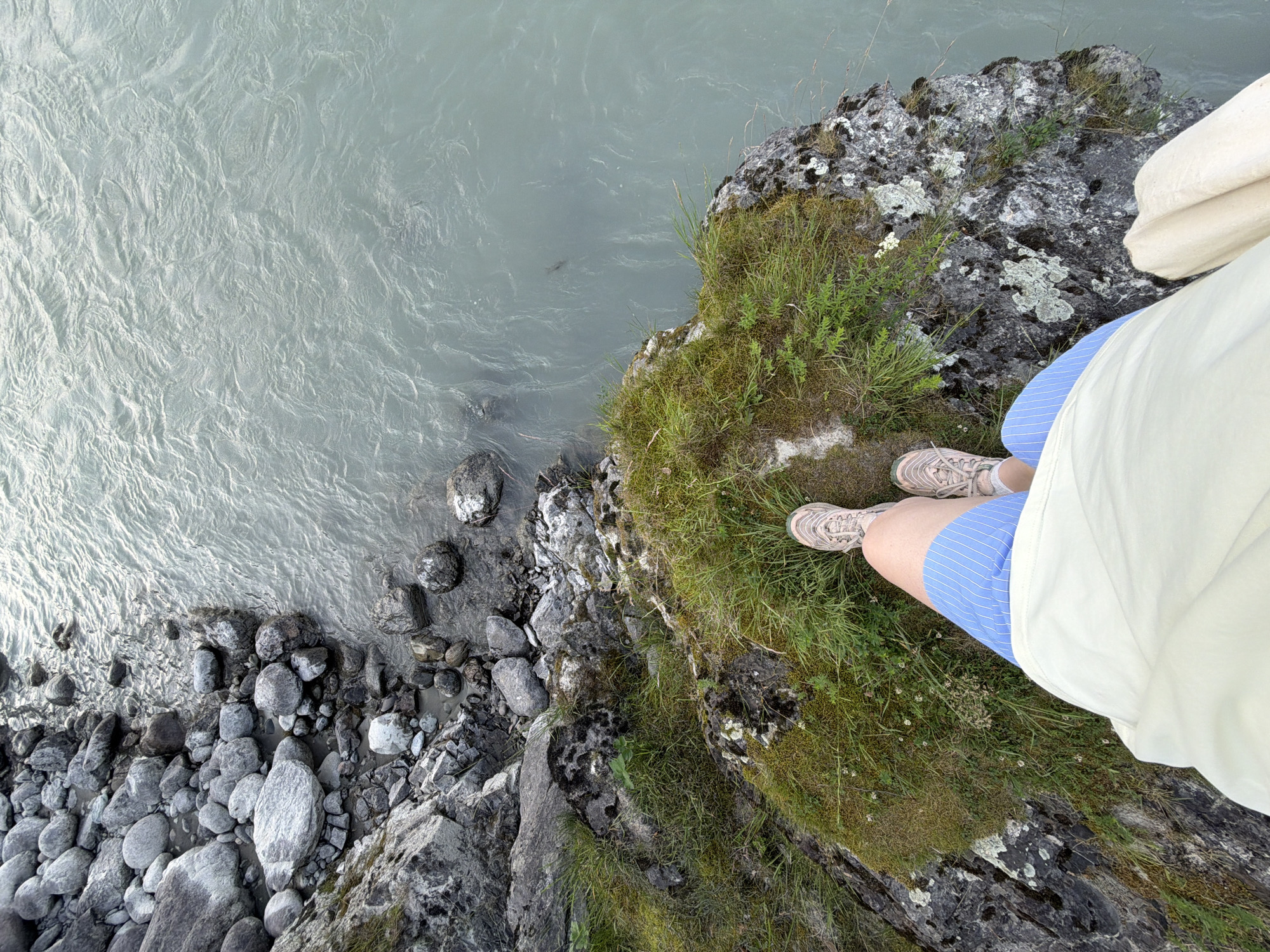 The image depicts a first-person perspective looking down at a person's legs and feet standing on a rocky, moss-covered outcrop next to a body of water. The person is wearing blue and white striped shorts and athletic shoes. The water appears to be a river with a light blue-green hue and visible ripples. The background consists of a rocky shoreline with large grey boulders and a grassy embankment. The location is not identified as Uznezya, Russia; no specific geographical or urban features are visible to confirm this.
