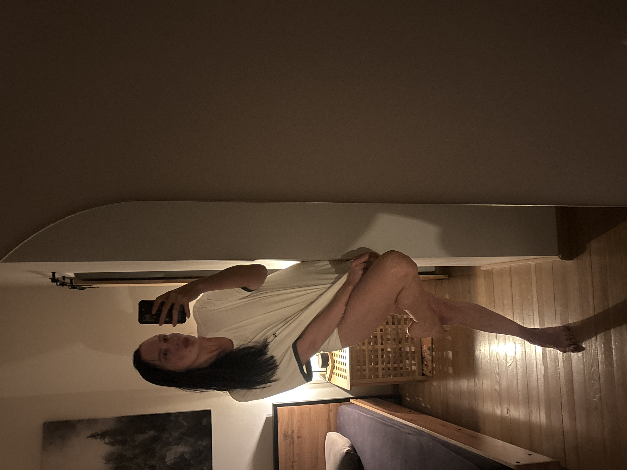 A woman is reflected in a mirror in a room. The room contains a bed with a purple headboard, a wooden bedside table, and a wooden floor. A painting is on the wall. A wicker basket is also visible. It is not possible to verify the location in Chaikovsky, Russia, from the image.