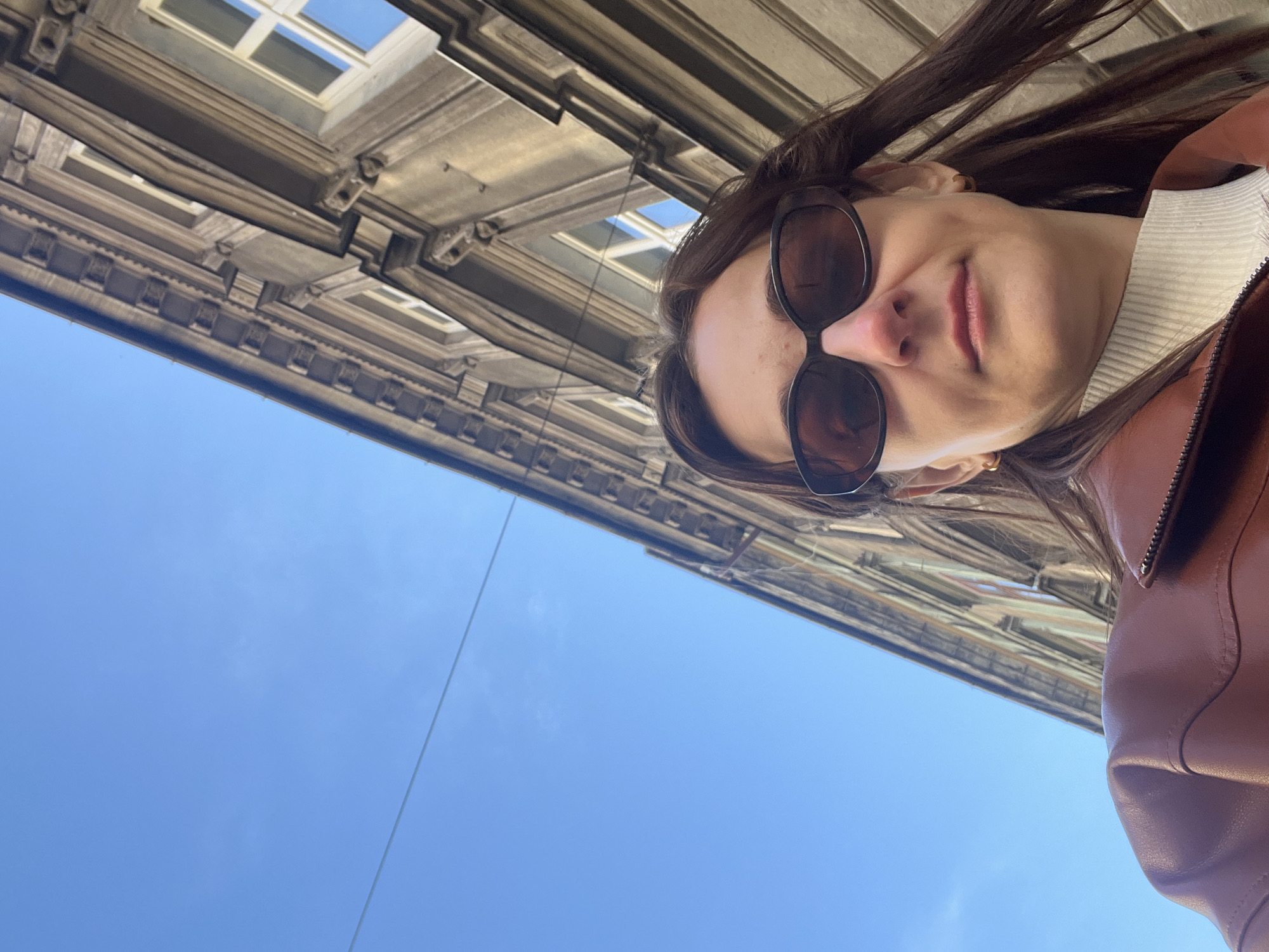 A low-angle shot looking upward captures a woman in sunglasses, with a building and clear blue sky in the background. The woman is wearing a brown leather jacket and a white turtleneck shirt. The building features rows of windows with multiple panes, set within a light-colored, ornamented facade. The sky is a cloudless, clear blue. The location is Unknown, Unknown.