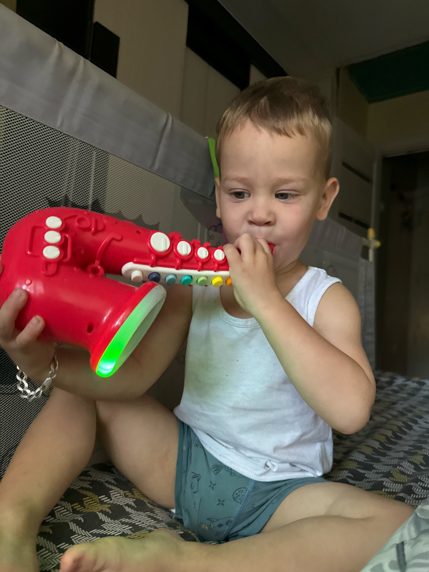 A young boy with blond hair, wearing a white tank top and blue shorts with a playful pattern, sits on a patterned blanket in what appears to be a room in Rusanovo, Russia. He is holding a bright red toy saxophone with colorful buttons and a glowing green opening. The boy is actively playing the saxophone, with his mouth near the mouthpiece and his hands holding the instrument. His gaze is directed downwards, suggesting focus on his play.
The setting includes a gray, mesh-sided crib or playpen surrounding the boy, with a darker grey padded railing at the top. The background is dimly lit, with glimpses of dark walls and some green and yellow elements suggesting the interior of a room. The lighting is somewhat dim, with the toy saxophone being a prominent source of color and light, particularly the green glow at its bell. The time of day is unclear, but the indoor setting and lighting suggest it is not bright daylight. The boy appears engaged and content with his toy. No visible text is present in the image.