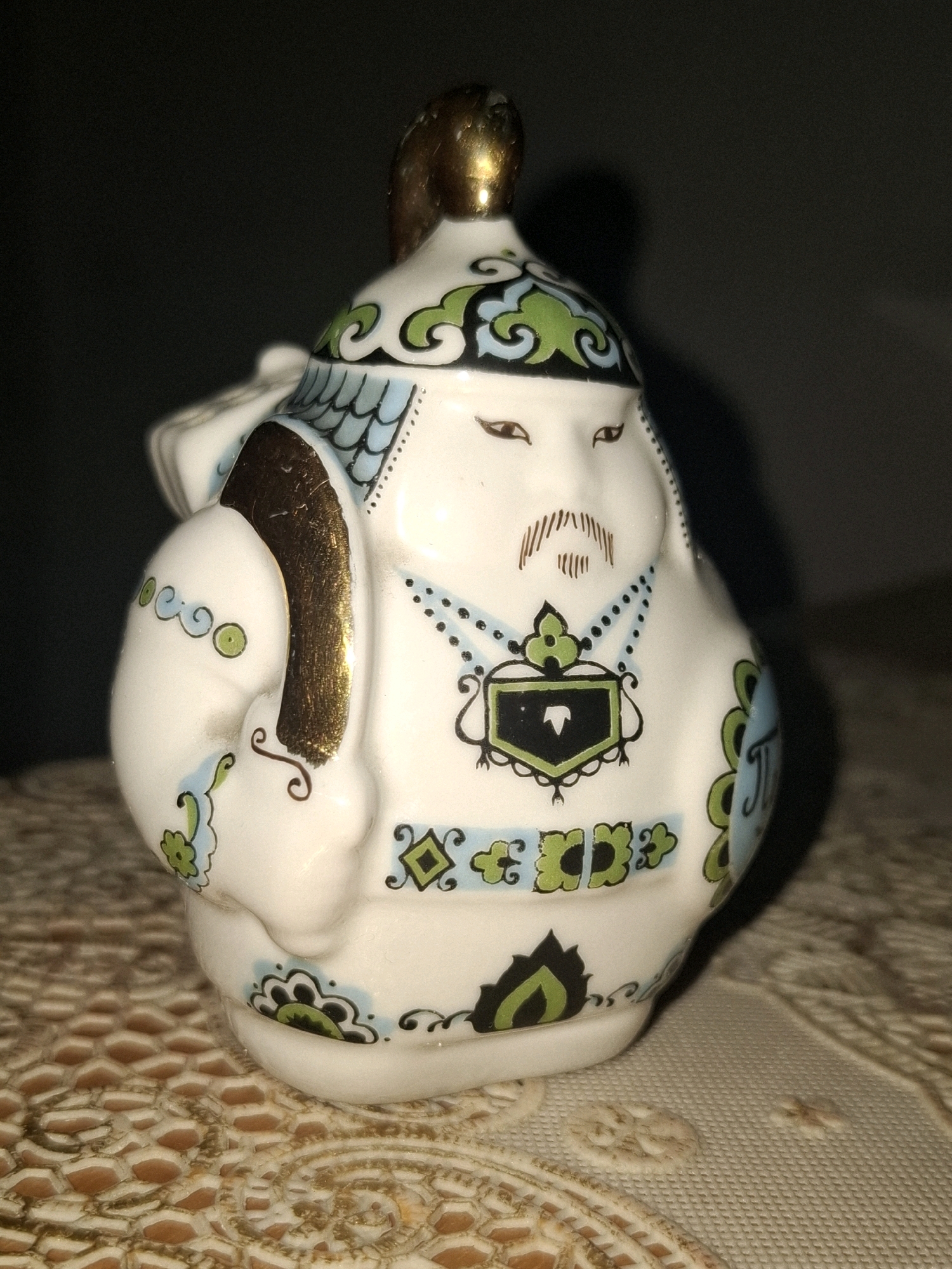 This is a close-up, slightly blurry photograph of a decorative ceramic figurine. The figurine is white with intricate blue, green, and gold detailing. It depicts a stylized, rotund figure with a cap topped with a gold adornment. The figure has painted facial features, including eyes, a mustache, and a beard. It is wearing ornate clothing with geometric and floral patterns. The figurine is positioned on a textured surface, possibly a doily or tablecloth, with a dark, out-of-focus background. The lighting is dim, casting a shadow to the right of the figurine. There is no indication of the city of Almaty, Kazakhstan, or any specific location within it. The image appears to be a still-life shot of the object.