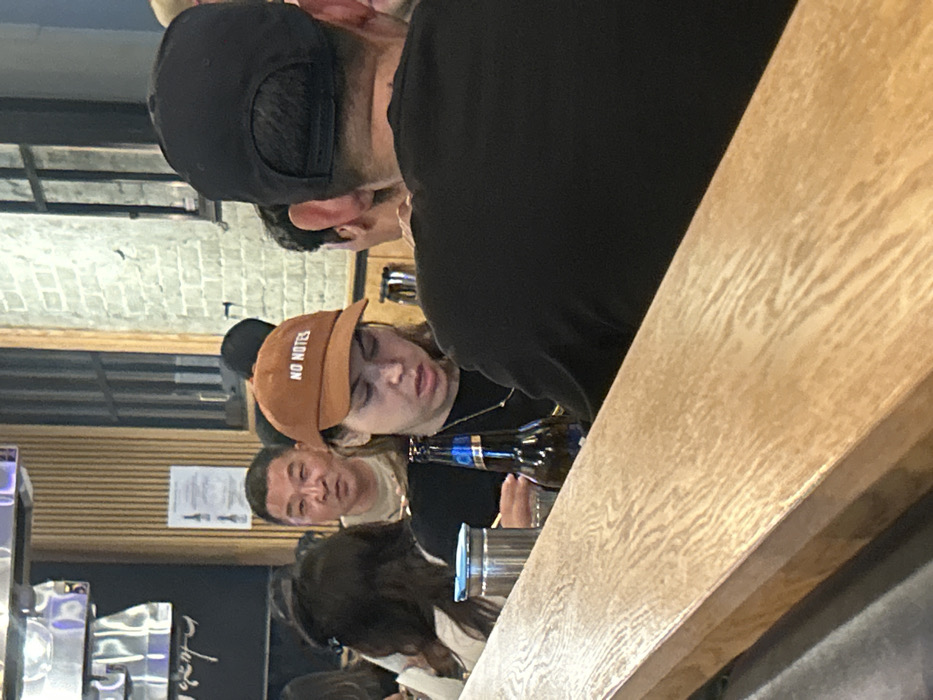 A woman wearing a "NO NOTES" baseball cap sits at a bar, eyes closed. She's holding a beer bottle. Beside her, a man is looking slightly to the left of the frame. In front of her, another woman is focused on something out of view. The scene is inside what appears to be a restaurant or bar, with a wooden countertop in the foreground. A black-clad man's shoulder and head are partially in frame at the top, and a window and menu are behind them.