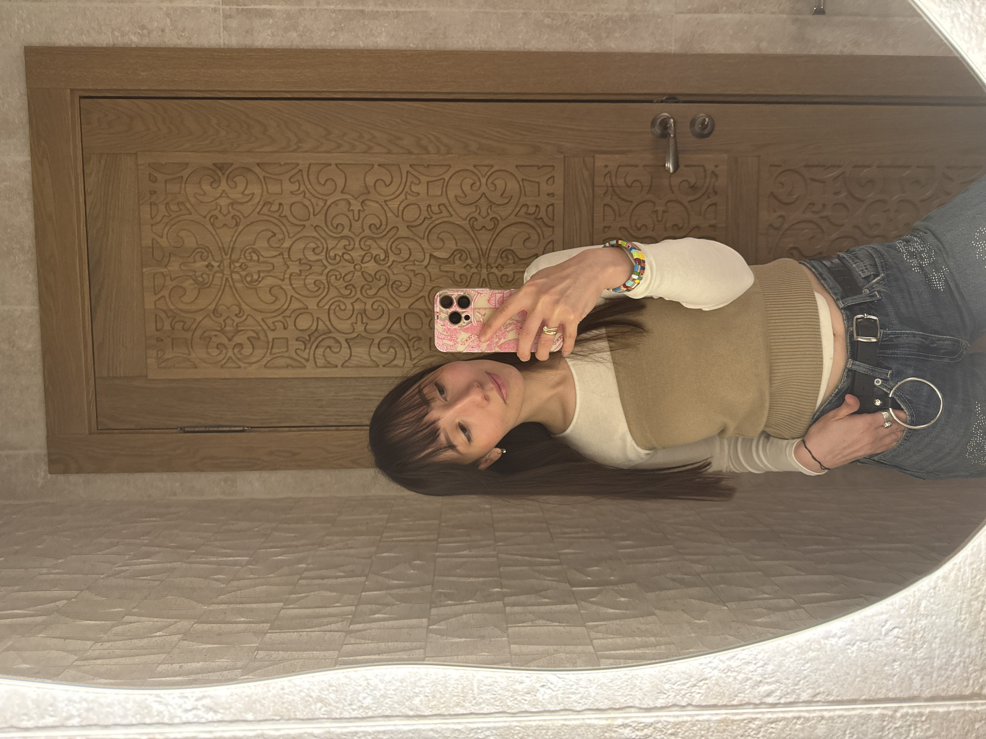 A young woman is taking a mirror selfie in a bathroom. The bathroom features a carved wooden door with a silver handle, and a wall with textured, light-colored tiles. The mirror is circular and has a light around the edge. The woman is wearing a white long-sleeved shirt, a beige vest, and blue jeans with silver embellishments and a belt. She is holding a pink phone with her right hand and has a colorful bracelet on her wrist. Her hair is dark and she has bangs. The location of the bathroom is unknown.
