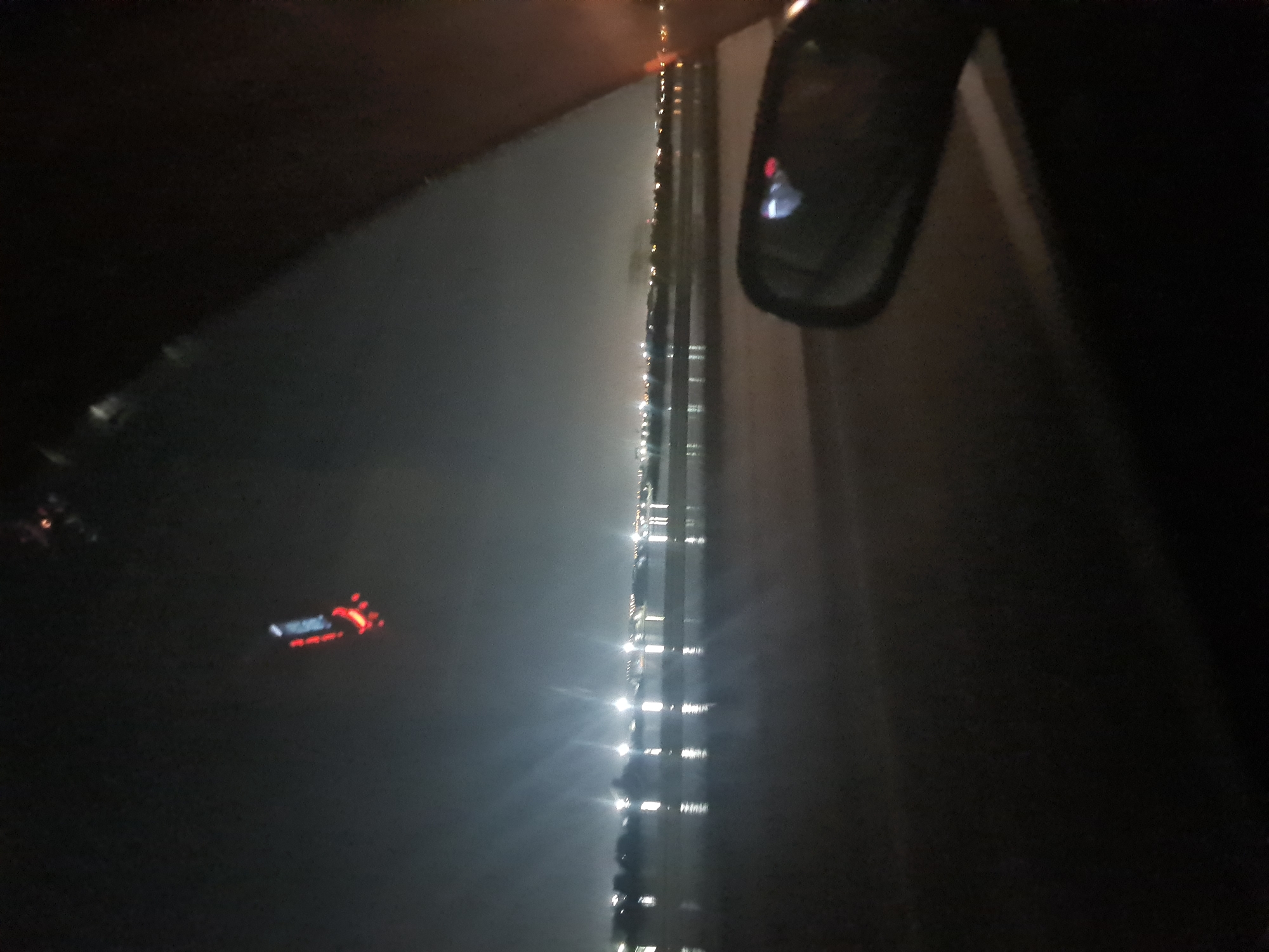 The image is a dark, nighttime photograph taken from inside a vehicle, likely a car, looking out a window. In the upper center of the frame, a small object with red lights is hanging from the ceiling of the vehicle. The view outside the window shows a body of water, possibly a river or lake, with numerous lights reflecting on its surface. In the distance, there is a faint outline of a city skyline with more lights, suggesting an urban area. A side-view mirror of the vehicle is visible in the lower right portion of the image. The scene appears to be on the outskirts of Irkutsk, Russia, near the Angara River, as suggested by the presence of the body of water and the city lights.