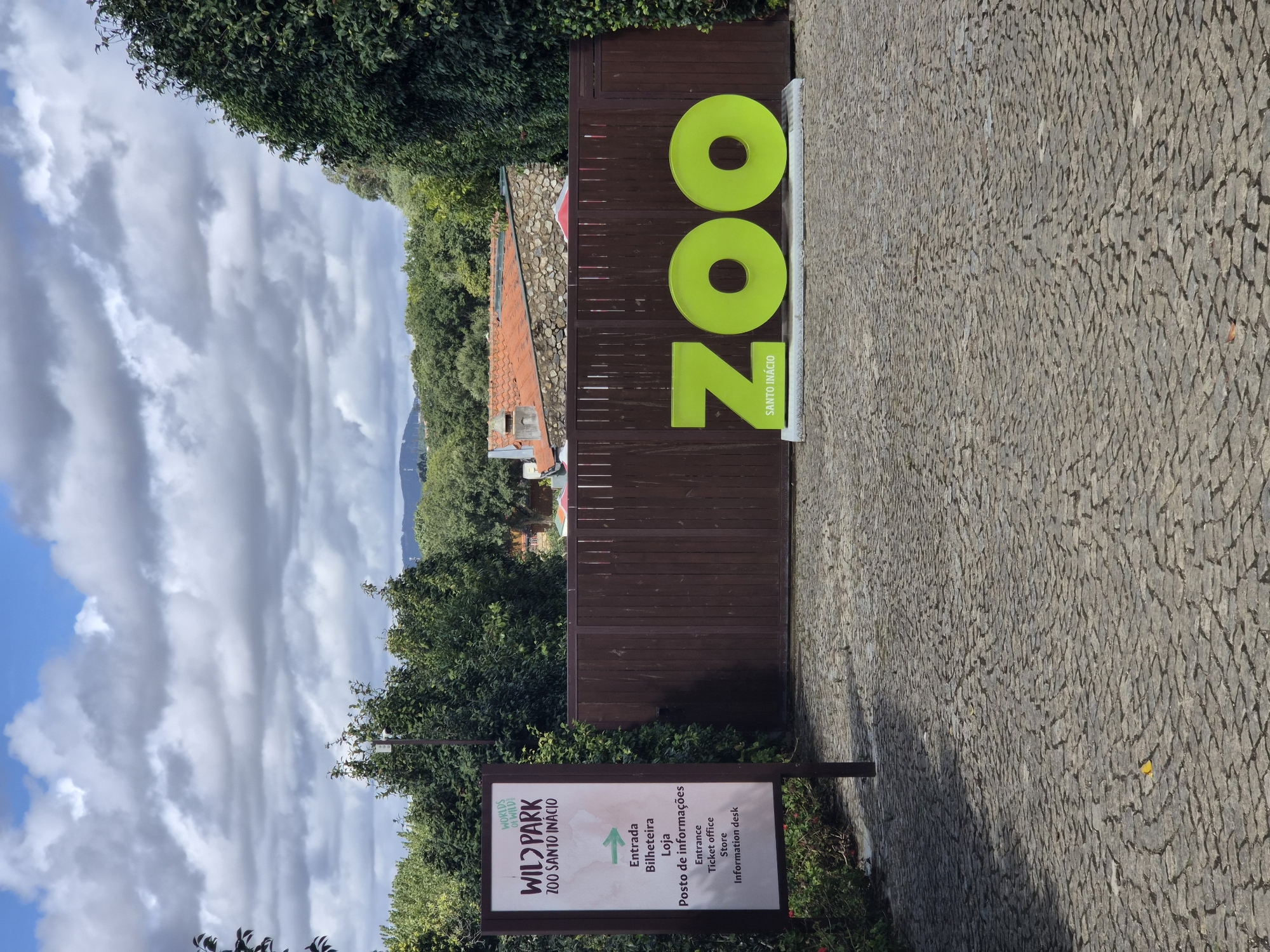 The image shows the entrance to the Zoo Santo Inácio, located in Vila Nova de Gaia, Portugal. A tall, narrow sign on the left displays "WILD PARK ZOO SANTO INÁCIO" in large font, with smaller text below listing "Entrada Bilheteira Loja Posto de informações" and its English translation: "Entrance Ticket office Store Information desk". To the right of the sign, a large, bright green "ZOO" logo is mounted on a dark wooden fence. The letters are three-dimensional, with the 'O's being large circles. The ground in front of the fence is paved with grey cobblestones arranged in a circular pattern. Behind the fence, there are glimpses of red-tiled roofs and lush green trees, suggesting a natural or park-like setting. The sky above is partly cloudy, with large, white clouds against a blue background. The overall impression is an outdoor entrance to a zoo on a slightly overcast day.