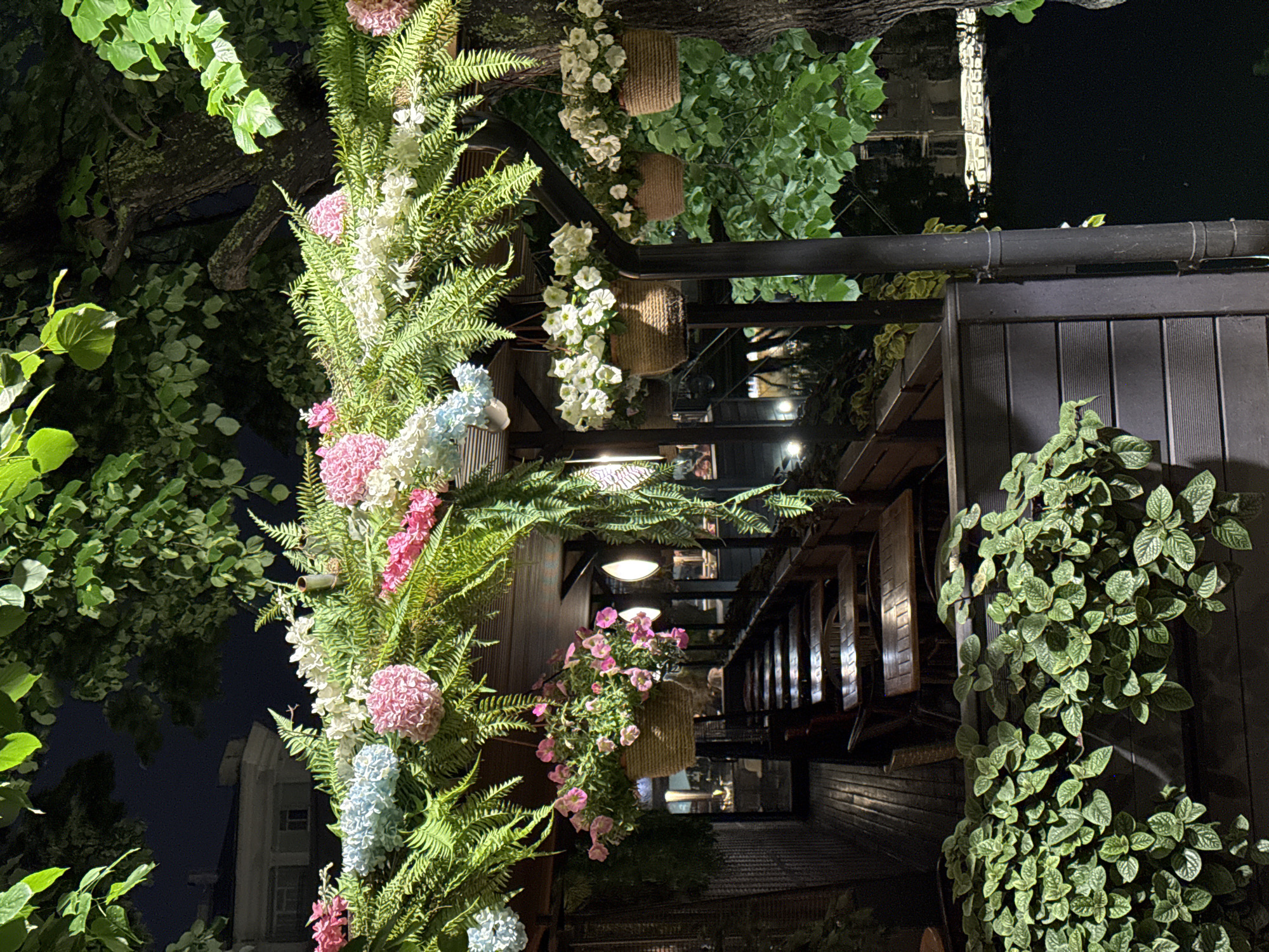 The image depicts an outdoor setting in Moscow, Russia, at night. Lush greenery surrounds a building, including ferns, climbing plants, and flowering arrangements with pink, white, and blue blossoms. Hanging planters with white flowers are visible. The building's exterior is constructed with dark wood paneling and metal framework. Interior lighting suggests a commercial establishment, possibly a restaurant, with tables and chairs visible through openings in the structure. A building can be seen in the dark sky.