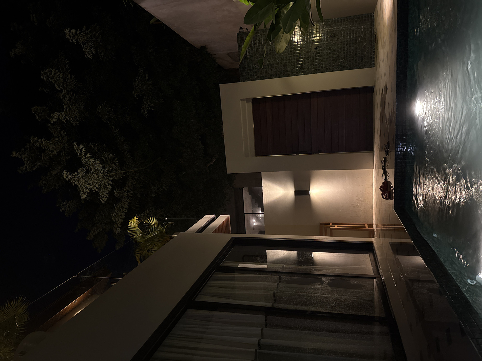 The image captures an exterior view of a building located in Kuta Selatan, Indonesia, at night. A rectangular pool with rippling water occupies the right side of the frame, reflecting light. Adjacent to the pool is a white-walled structure with dark brown wood paneling. The building features large windows with visible curtains inside. The area is illuminated by outdoor lighting, highlighting the architecture. Dense vegetation, including trees and plants, is visible on the left side of the image, contrasting with the modern architecture.