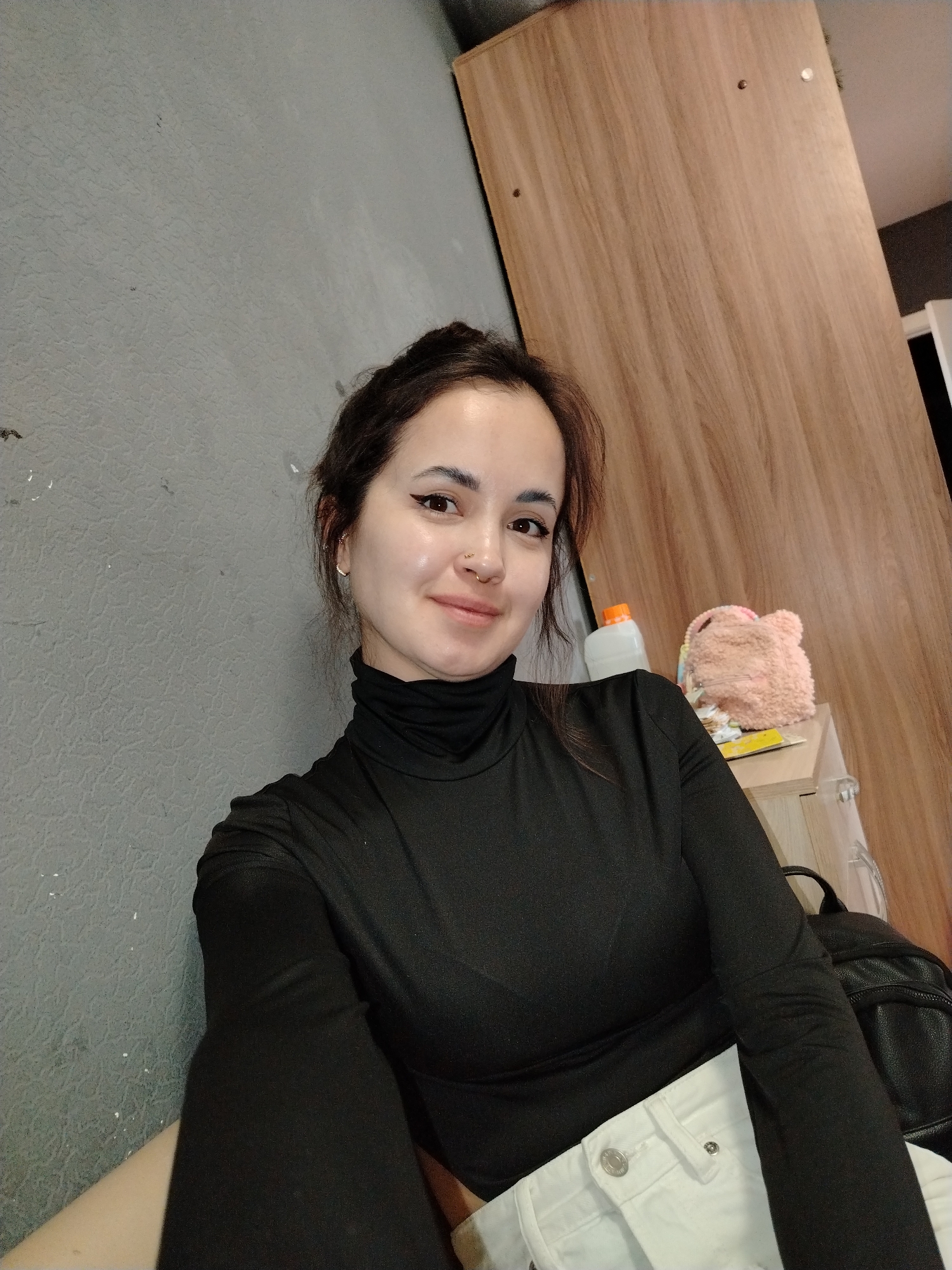 A woman is shown in a medium close-up shot. She has dark hair and is wearing a black turtleneck shirt and white pants. She is smiling slightly. Behind her is a gray wall on the left, and a wooden cabinet or door on the right. On top of a cabinet or drawer, there are a white plastic bottle, a pink furry bag and a book. A black backpack is beside her. It is not possible to confirm the location in the city of Noviy, Russia with the given image.
