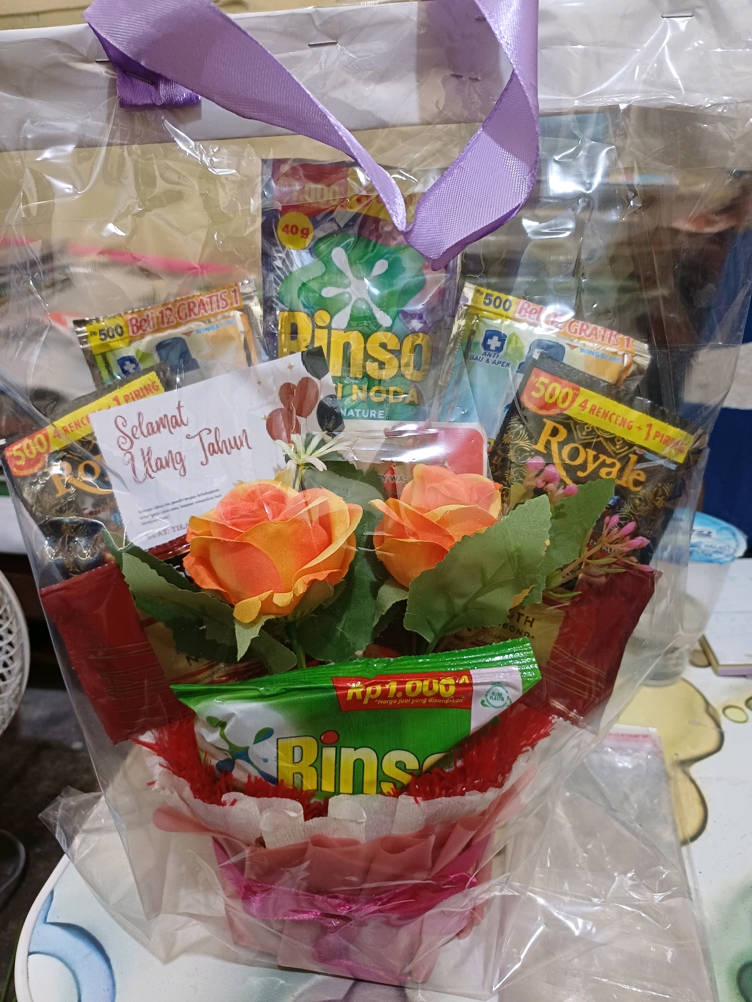 The image shows a gift basket that appears to be for a birthday. The basket is made of various items including laundry detergent packets, flower arrangements, and a greeting card. The greeting card has "Selamat Ulang Tahun" written on it, which translates to "Happy Birthday" in Indonesian. The basket is wrapped in clear plastic and tied with a purple ribbon. The background suggests an indoor setting, possibly a home or a shop, with hints of furniture and other objects that are not clearly identifiable. The scene does not provide any specific visual cues that would allow for precise location identification within Dumai City, Indonesia, or any other specific geographical location.