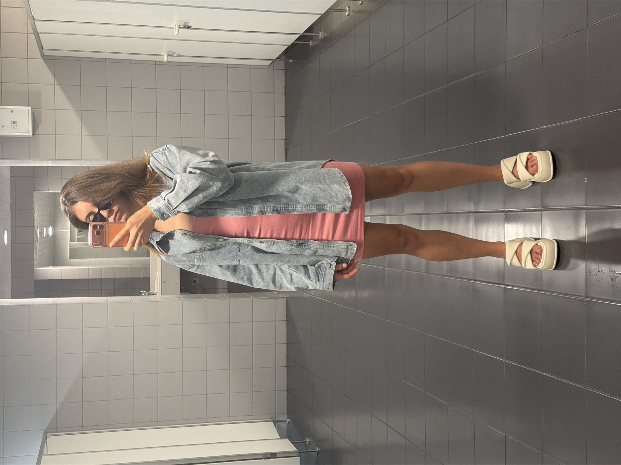 The image, taken in Moscow, Russia, depicts a young woman capturing a mirror selfie in what appears to be a public restroom or changing facility. The original photograph is oriented sideways, with the woman standing horizontally across the frame.
The woman has long, light brown hair and is wearing dark sunglasses. She holds a smartphone, encased in a light orange or coral protective cover, up to her face to take the picture, partially obscuring her expression. Her attire includes a sleeveless, light pink or coral dress, layered with a light blue, oversized denim jacket. On her feet, she wears cream-colored, puffy-strapped flat sandals.
The setting is characterized by utilitarian tiled surfaces. The floor is composed of large, dark gray square tiles, while the walls are covered in smaller, light gray or white tiles. In the background, white partition doors, likely for toilet stalls or changing cubicles, are visible. The lighting is bright and artificial, typical of an indoor public space. There is a large mirror reflecting parts of the room. No readable text is visible on any surfaces or objects in the image. The time of day is not discernible from the indoor lighting.