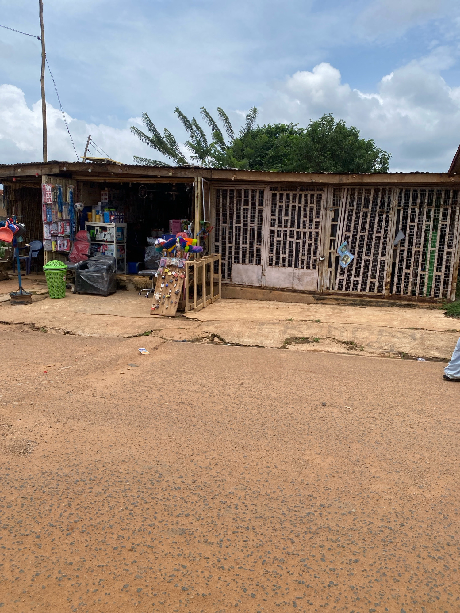 The image shows a roadside shop in Iworoko, Nigeria. The shop has a corrugated metal roof and a front made of metal grilles. Various items are displayed for sale, including locks on a wooden stand, cleaning supplies, and other household goods. A green trash can and a wrapped chair are also visible outside the shop. The ground in front of the shop is a mix of concrete and dirt, with the road in the foreground appearing to be asphalt. The sky is cloudy, and trees are visible in the background.