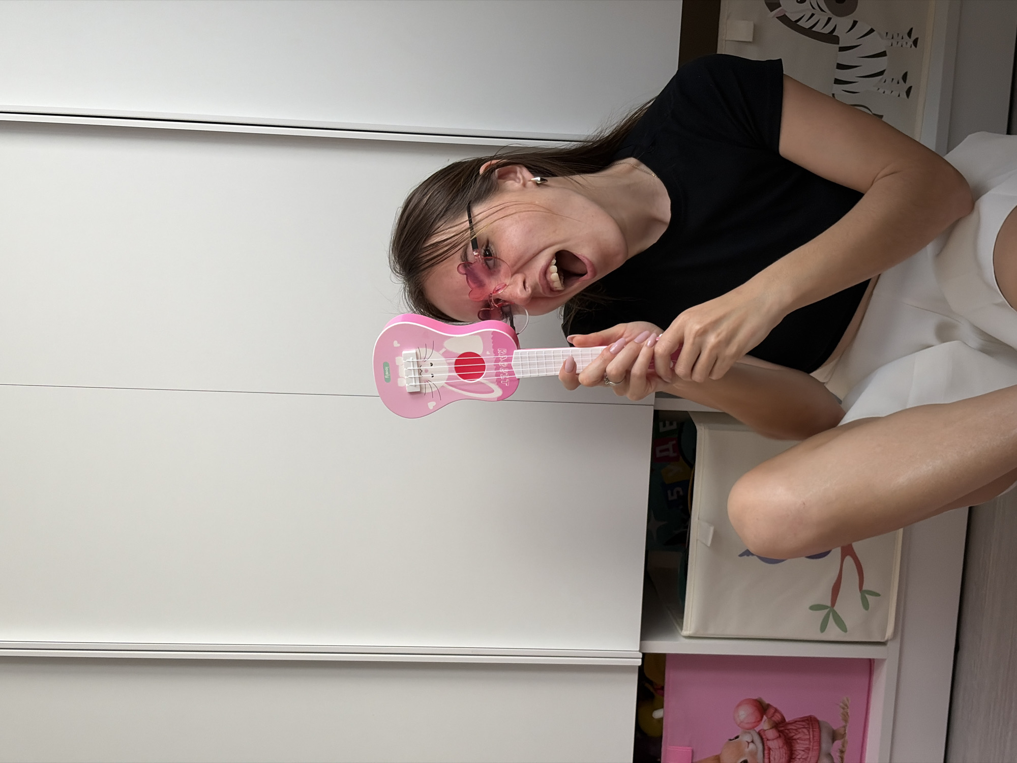 A woman with dark hair and glasses is holding a pink toy guitar. She is wearing a black top and white shorts. She is indoors. The background includes white closet doors and storage boxes with designs on them. The location is Izhevsk, Russia.