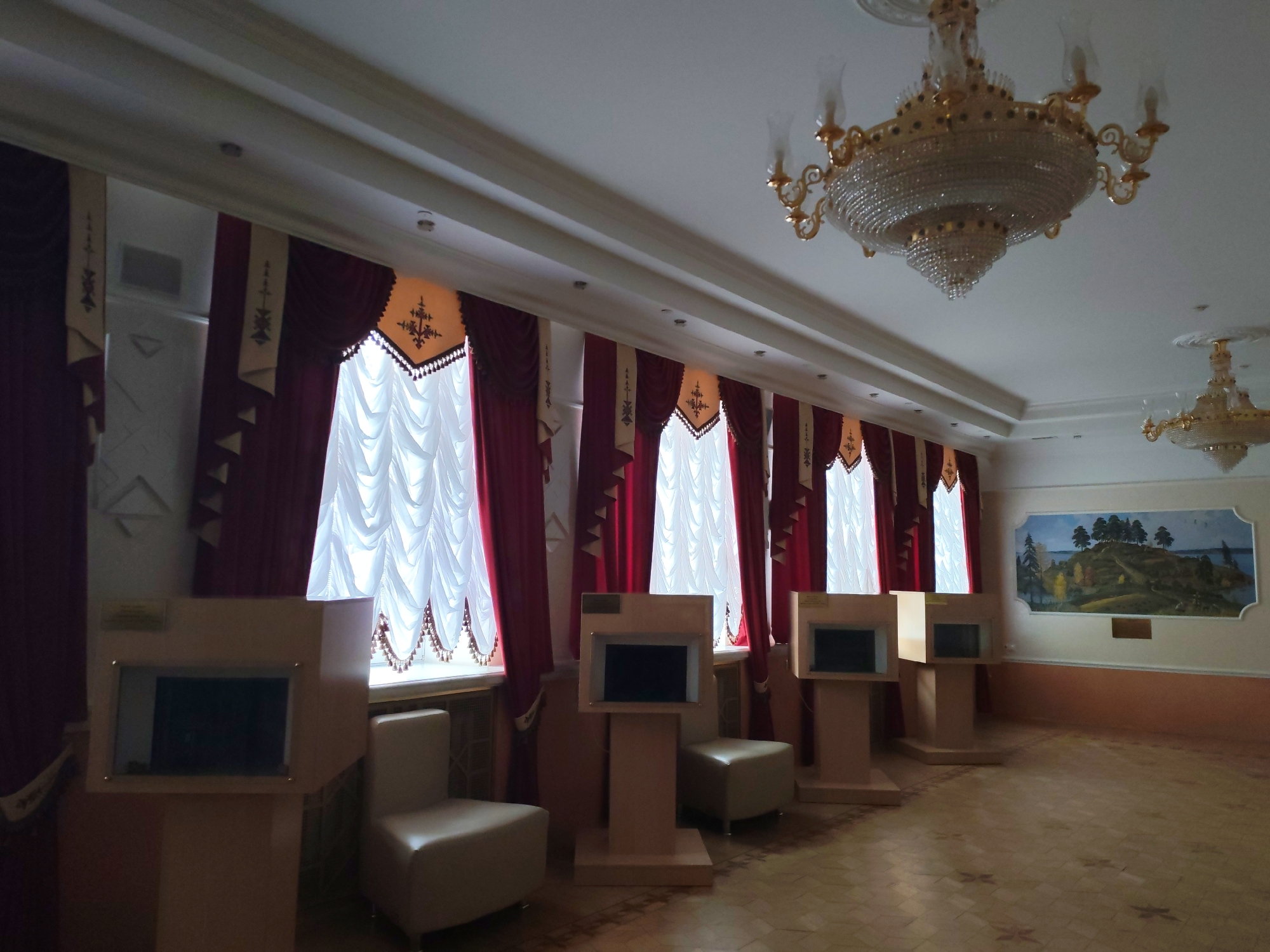 This media file presents a spacious, well-lit interior room, likely a hall, gallery, or exhibition space within a public building in Izhevsk, Russia.
The primary subjects are the architectural and decorative elements of the room. A series of tall, rectangular windows line one wall, adorned with opulent deep red velvet curtains and white sheer inner curtains. The red curtains feature elaborate valances with intricate golden-beige embroidered patterns and a scalloped bottom edge, suggesting a formal or traditional design.
From the white, molded ceiling hang two ornate chandeliers with multiple tiers of crystals and golden metal frames, providing artificial illumination alongside recessed ceiling lights.
Along the windowed wall, five light-colored wooden or laminate display kiosks are arranged. Each kiosk contains a dark, rectangular screen, likely an interactive display. Small, light-colored upholstered chairs or ottomans are placed in front of some kiosks, indicating areas for visitors to sit and engage with the content.
On the opposite wall, a large framed painting or mural depicts a serene landscape scene featuring a body of water, a tree-covered hill, and a distant horizon. The floor is covered with patterned tiles, possibly marble or ceramic, with subtle floral or geometric motifs.
No people are visible in the image. The arrangement of kiosks suggests an informational or exhibition purpose, perhaps in a museum, cultural center, or a visitor's lounge. The natural light filtering through the windows indicates it is daytime. No specific text is legible on the small plaques attached to the kiosks or below the painting. The overall ambiance is grand and inviting.