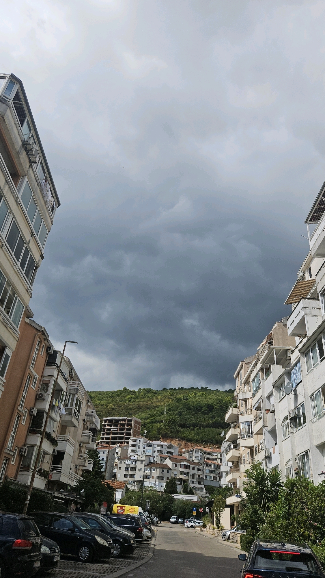 The image depicts a street scene in a coastal city, likely in the Mediterranean, with apartment buildings lining both sides of a narrow road. The buildings are a mix of white, beige, and terracotta, with numerous balconies and windows. Some buildings appear older, while others are more modern, and an unfinished concrete structure is visible on a hillside. The street is lined with parked cars, predominantly dark-colored sedans and SUVs. Lush green vegetation covers the hillsides in the background, contrasting with the overcast, grey sky. A street lamp is visible on the left side of the road. The overall impression is of a densely populated urban area nestled against a natural landscape.