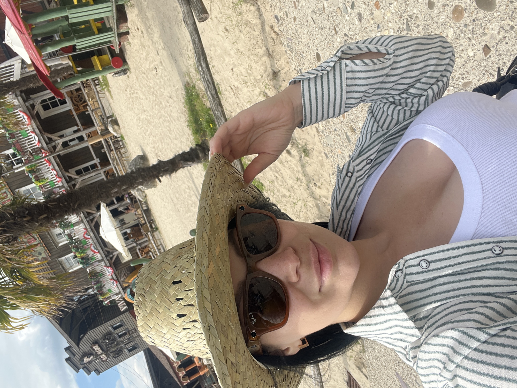 In a selfie taken at a Wild West-themed attraction in Collendoorn, Netherlands, a woman with dark hair wears a straw cowboy hat and sunglasses. She has on a white tank top and a striped button-down shirt, partially unbuttoned to reveal her décolletage. Her right hand adjusts her hat. The background features wooden buildings with balconies decorated with flags, a palm tree, sandy ground, and some playground equipment. The sky is partly cloudy.