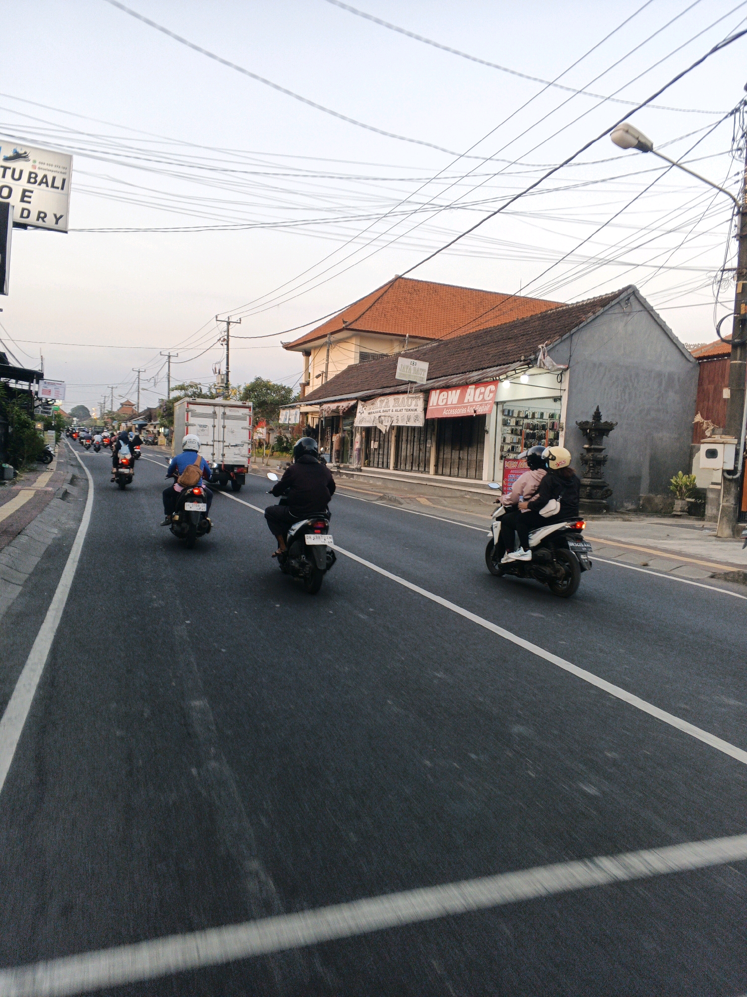 This image depicts a street scene in Denpasar, Indonesia, likely during the early morning or late afternoon, given the soft lighting. The foreground is dominated by a road with visible white lane markings. Several motorcycles are present, with riders and passengers wearing helmets, indicating adherence to traffic regulations. One motorcycle in the middle ground has a rider in a blue shirt and a passenger with a light-colored bag on their back. Further ahead, another motorcycle with two occupants, one in a dark jacket and the other in a pink hoodie, is also visible. In the distance, a white truck is traveling on the road. The background features buildings lining the street, including one with a red-tiled roof and signage for "New Acc Accessories & Repair." Overhead, a complex network of electrical wires crisscrosses the sky, a common sight in urban areas. A street lamp is mounted on a pole on the right side of the image. The overall atmosphere suggests an active but ordinary day in a bustling Indonesian city.