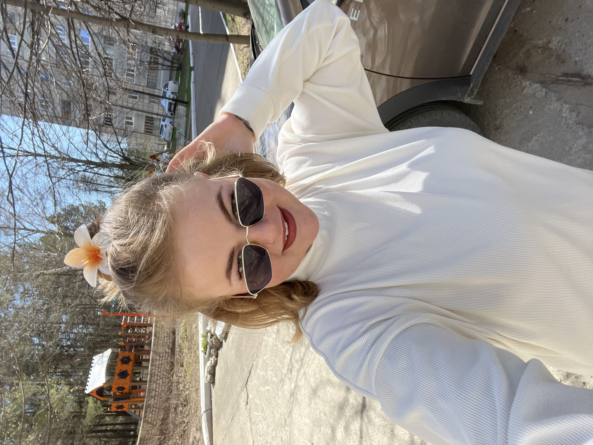 The image shows a woman wearing sunglasses and a white turtleneck sweater standing outdoors next to a car. There is a flower in her hair. Behind her is a playground, a building, and trees. The location is likely in Chaikovsky, Russia, based on the provided information.