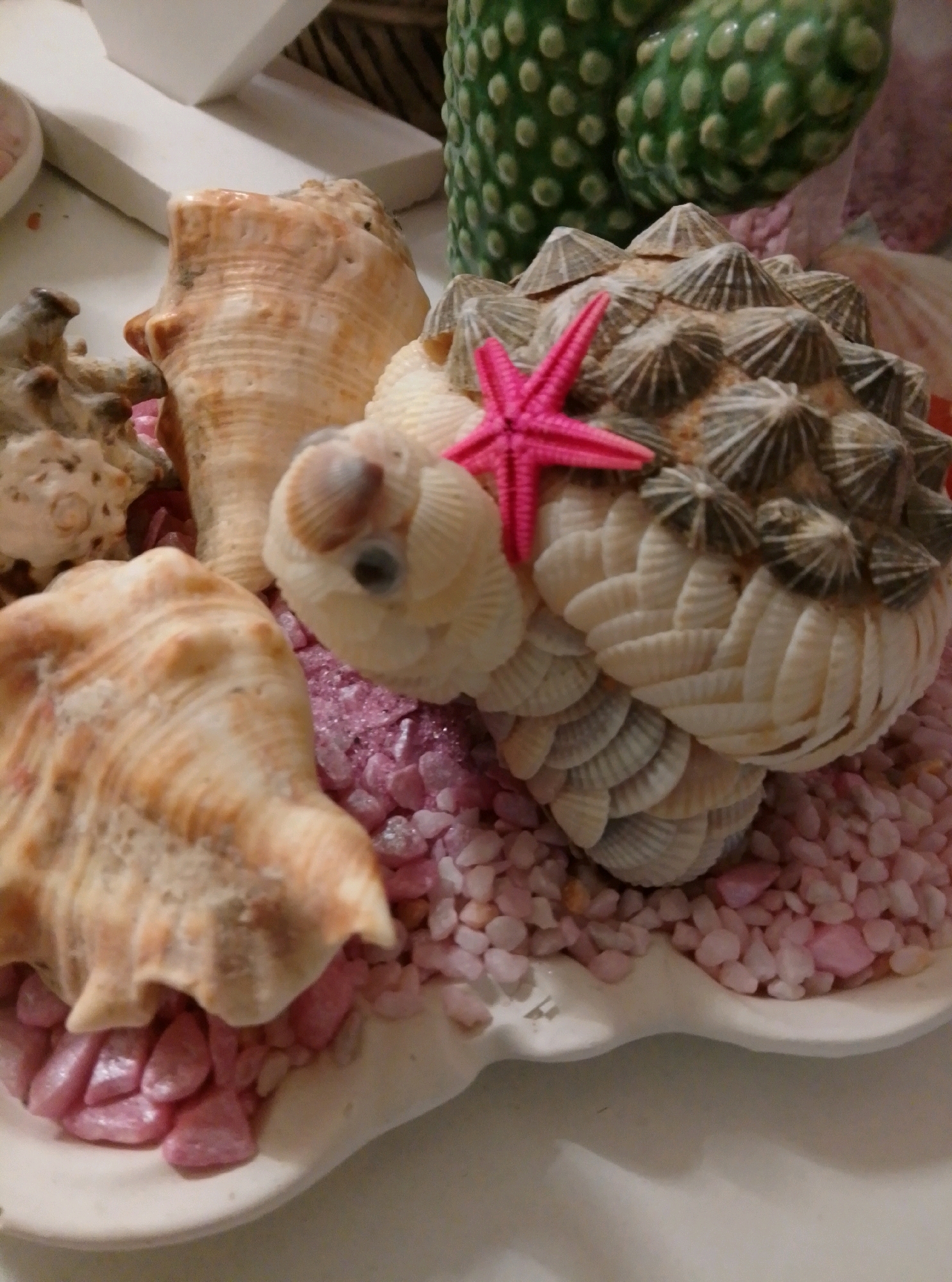 This image shows a decorative seashell arrangement on a white scalloped platter. The arrangement features several large conch shells, smaller shells that form a textured ball shape, and a small pink starfish. The platter is filled with small, pink, crushed stones. In the background, a green, bulbous, textured object is visible, possibly a ceramic decoration. The scene appears to be indoors, on a light-colored surface. There is no indication of a specific location in the city of Wedel, Germany.