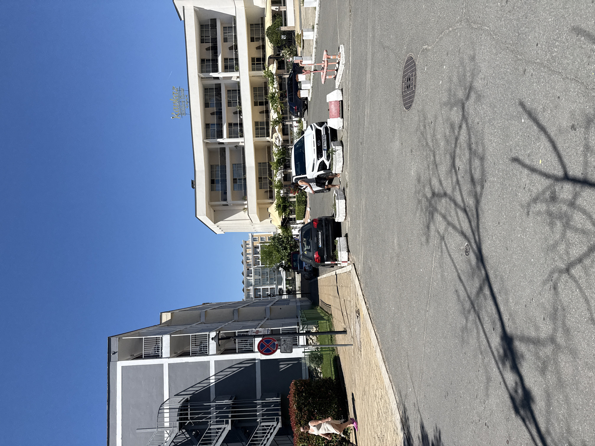 This is an eye-level, medium shot taken on a sunny day in Nesebar, Bulgaria, facing a street intersection. In the foreground, the asphalt road is visible with shadows of tree branches cast upon it, along with a manhole cover. To the left, a woman in a white dress and pink sandals walks on a cobblestone sidewalk next to a gray multi-story building with white trim and balconies. Adjacent to this building is a metal spiral staircase and a signpost with a red and blue circular no-parking symbol. In the background, on the right, a white multi-story hotel building with balconies and yellow awnings is situated, with a sign on top that reads "Kavkaz". Several cars are parked in the area, including a black Audi and a white Hyundai. Trees and bushes are also present, contributing to the urban outdoor environment.