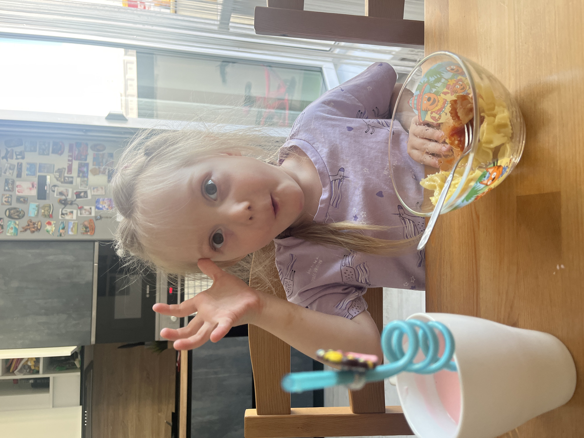 The image shows a young blonde girl with her hair in pigtails sitting at a wooden table in a kitchen. She is wearing a light purple t-shirt with unicorn and heart patterns. Her left hand is raised to her forehead, and she has an surprised expression with her mouth slightly open. In front of her is a clear glass bowl containing pasta with some small meat pieces, and a spoon. To her left is a white mug with a pink liquid and a blue swirly straw. The background shows kitchen cabinets and a window with vertical blinds. The image is taken from a medium shot, focusing on the girl and her meal. The provided crops highlight the girl's face, her hand, the meal, and the drink. The image is not geotagged and cannot be placed in Chelyabinsk, Russia.