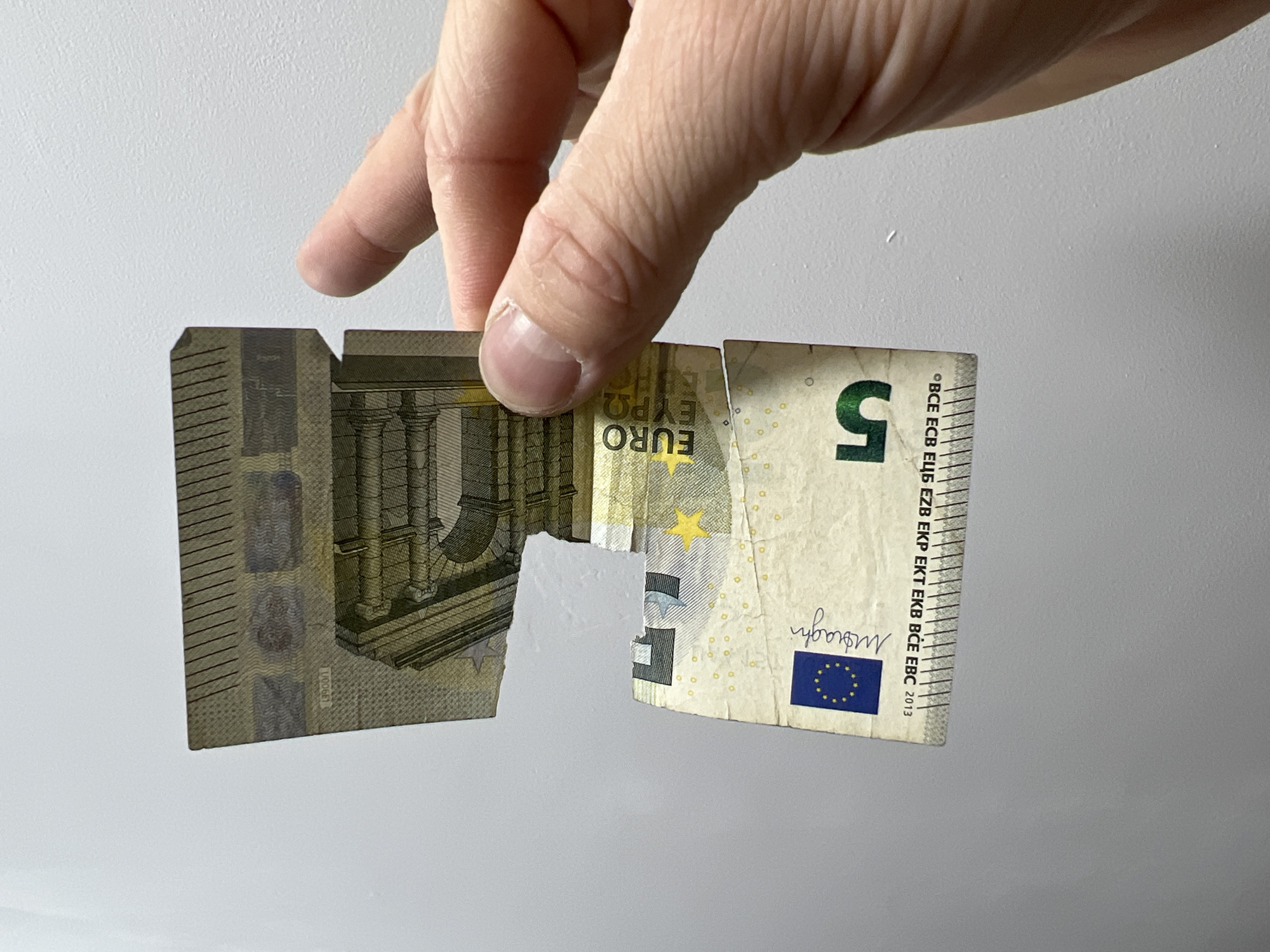 A partially torn 5 Euro bill is being held by a hand against a plain white wall. The bill is torn in the middle and the two halves are slightly separated, revealing the white wall in between. The hand is fair-skinned, with the thumb and index finger visible holding the two halves of the bill. Text on the bill includes "EURO", "Moraghi" and "5". A small number "0006E1" is also visible on the bill.