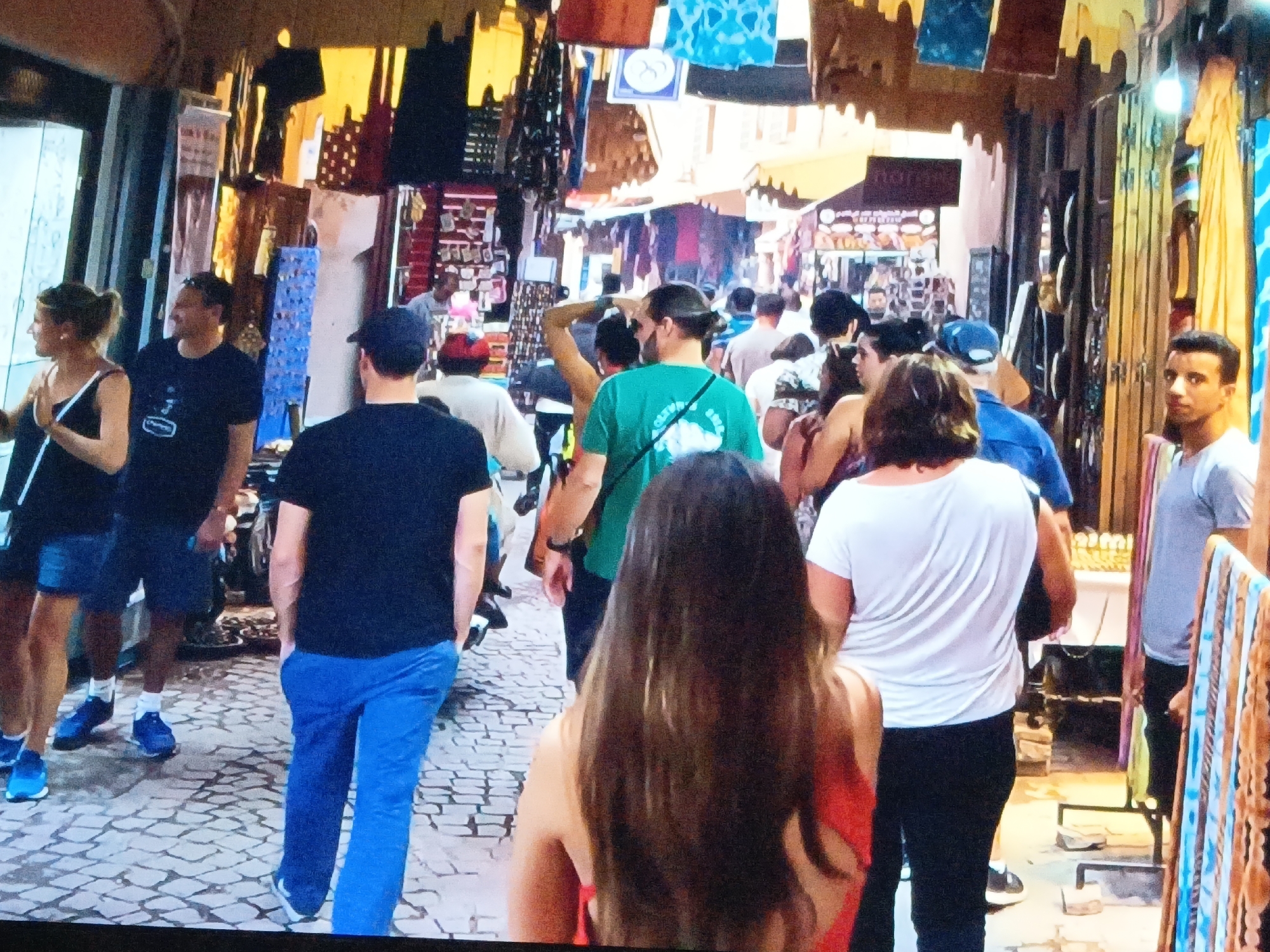 The image depicts a busy marketplace or bazaar, likely in a tourist area, with many people walking through a narrow street lined with shops. The scene is filled with vibrant colors from the merchandise hanging overhead and displayed in the stores. Shoppers of various ages and ethnicities are browsing and walking through the marketplace. A man in blue pants is the foreground. It is not in Juan Griego, Venezuela.