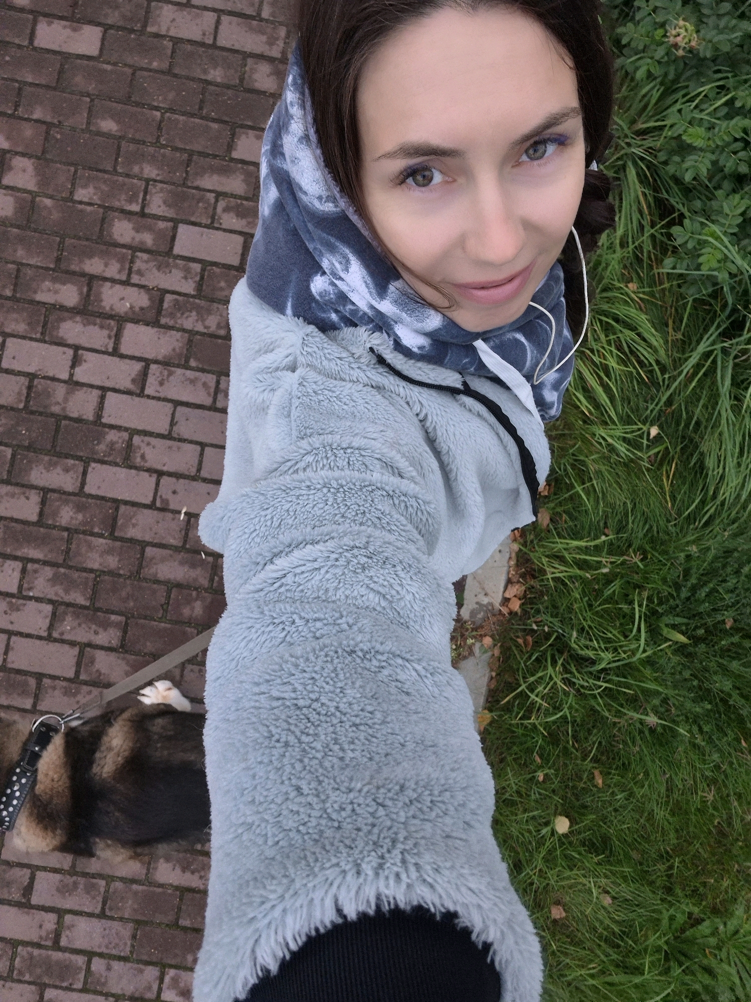 A high-angle selfie shows a woman with dark hair, wearing a light blue fuzzy jacket and a tie-dye hooded sweatshirt. She has headphones in her ears and is holding a dog on a leash. The dog is medium-sized with brown and black fur. They are standing on a brick pathway next to a grassy area. The scene appears to be outdoors, possibly in a park or residential area within the city of Chaikovsky, Russia. The brick pathway is laid out in a pattern, and the grass is lush and green. The woman's face is visible, looking towards the camera with a slight smile. Her makeup includes a touch of purple eyeliner. The overall lighting suggests it is daytime, possibly overcast.