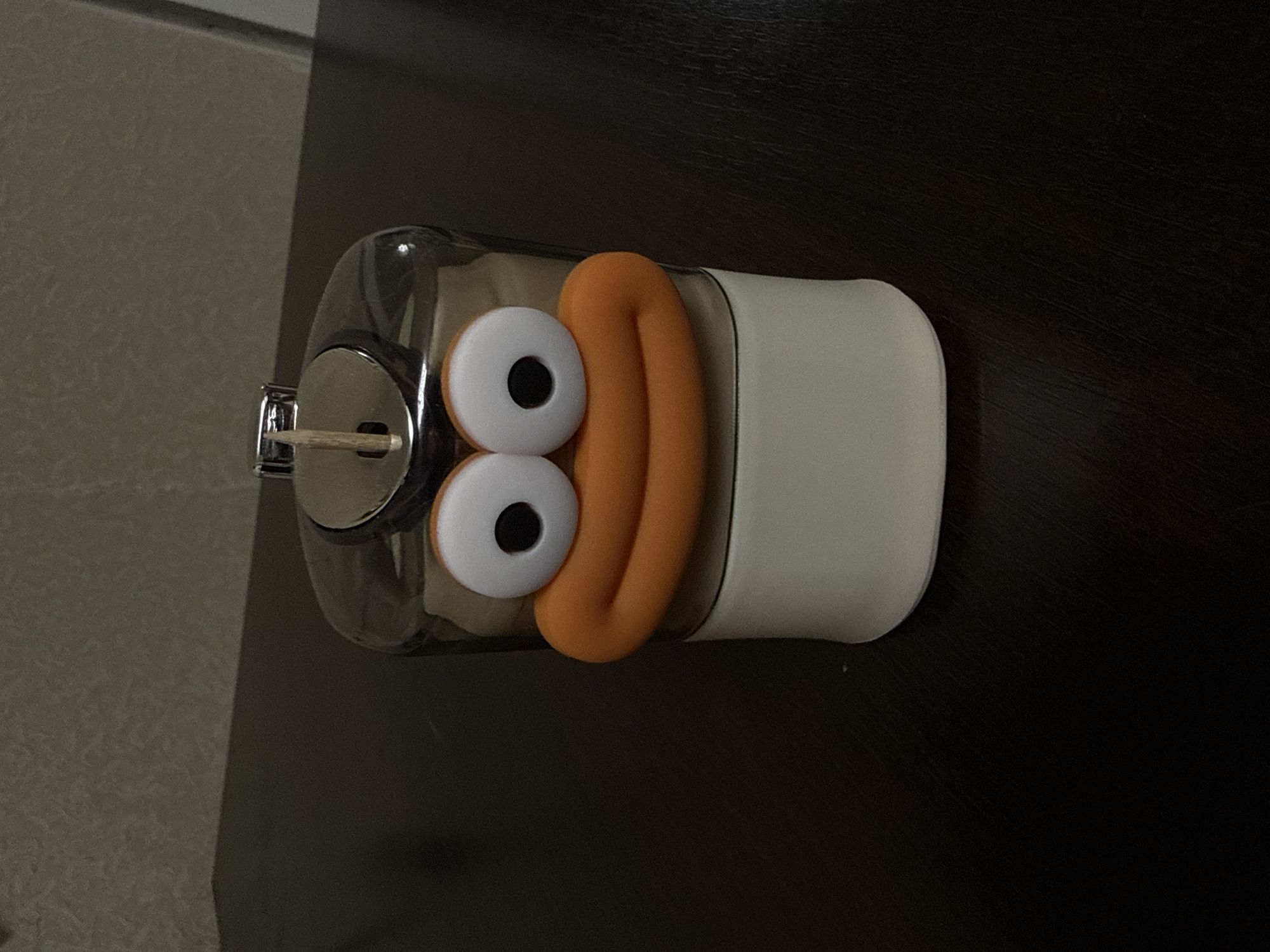 A whimsical, cartoonish toothpick dispenser sits on a dark, reflective surface. The dispenser is designed to resemble a character's face, with large, circular white eyes featuring black pupils and a prominent, bright orange, puckered mouth. Above the face, a clear plastic casing contains toothpicks, and a silver cap with a slot for dispensing them is visible. A single wooden toothpick protrudes from the dispensing slot. The dispenser has a white base. The background is dark and indistinct, suggesting an indoor setting. The location in Reppolovo, Russia, is not visually determinable from the image.