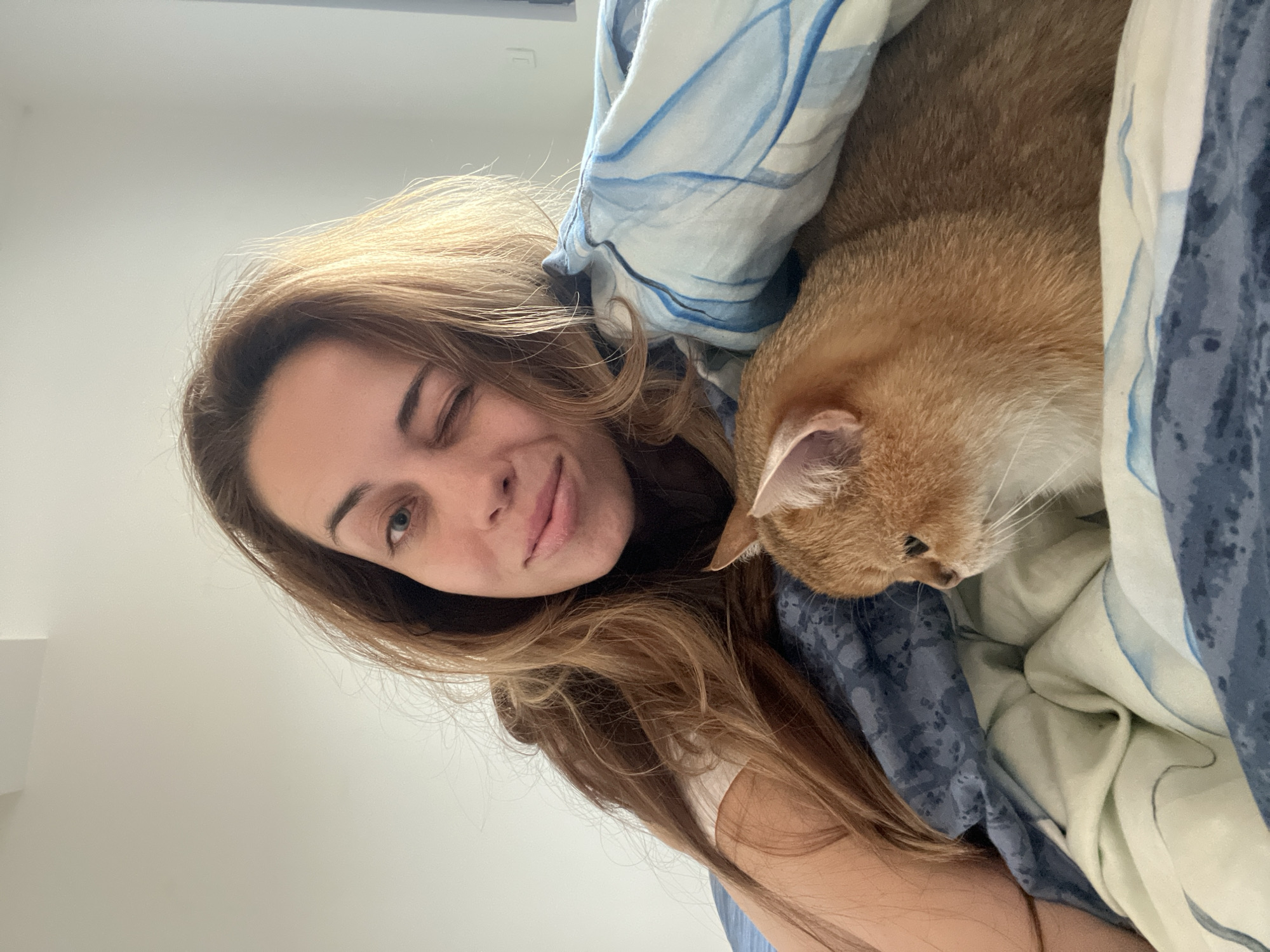 The image shows a woman and a cat in a bed with blue patterned bedding. The woman is winking and smiling at the camera. She has long, light brown hair. The cat is light orange in color and lying near the woman under the bedding. The environment appears to be a bedroom. There is no information to identify the location as Bjeliši, Montenegro.
