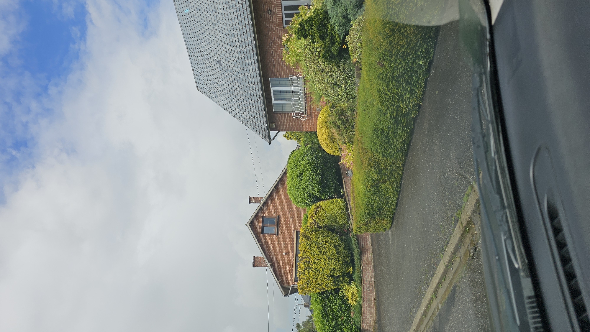 This image depicts a residential street scene in Thuin, Belgium, under a partly cloudy sky during daytime. The perspective suggests the photo was taken from inside a vehicle, with a dark interior panel partially visible along the right edge of the frame.
On the left side of the image, a dark, paved road or driveway is visible, running parallel to a low curb or a light-colored brick wall that separates it from the properties.
The right side of the scene is dominated by two brick houses surrounded by extensive, well-maintained landscaping. The house closer to the viewer features a reddish-brown brick facade, a dark grey slate roof composed of numerous small tiles, and a white-framed window with a decorative metal grate beneath it. Power lines stretch across the mid-ground.
Behind and slightly to the right of the first house, a second brick house is visible, also constructed of reddish-brown brick with a dark slate roof and two chimneys. The properties are adorned with a variety of lush shrubs and hedges, exhibiting different shades of green and vibrant yellow-green. A long, neatly trimmed green hedge runs along the property boundary adjacent to the road.
The sky in the upper left section of the image shows patches of bright blue amidst soft white and grey clouds. No people or specific activities are taking place, presenting a quiet, typical residential street view. There is no visible text in the image.