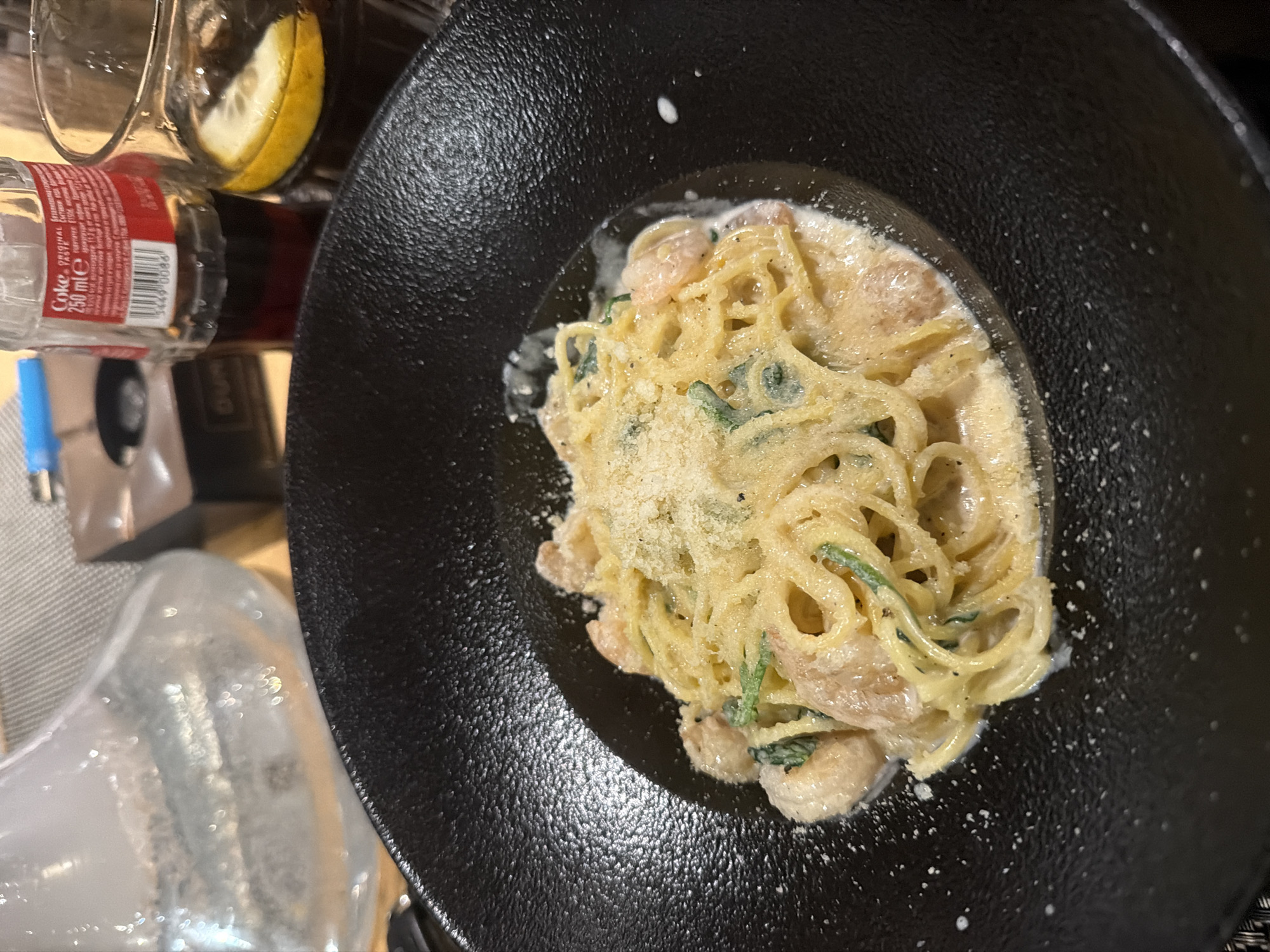 A top-down shot shows a plate of pasta in a black bowl. The pasta is a creamy color and appears to be covered in parmesan cheese, with visible green herbs mixed in. The black bowl has a wide rim and textured surface.
To the left of the plate are a variety of items, including a glass with a lemon slice, a small glass Coca-Cola bottle, and other indistinguishable items. The setting appears to be a restaurant table. There's no definitive proof the photo was taken in Plovdiv, Bulgaria.