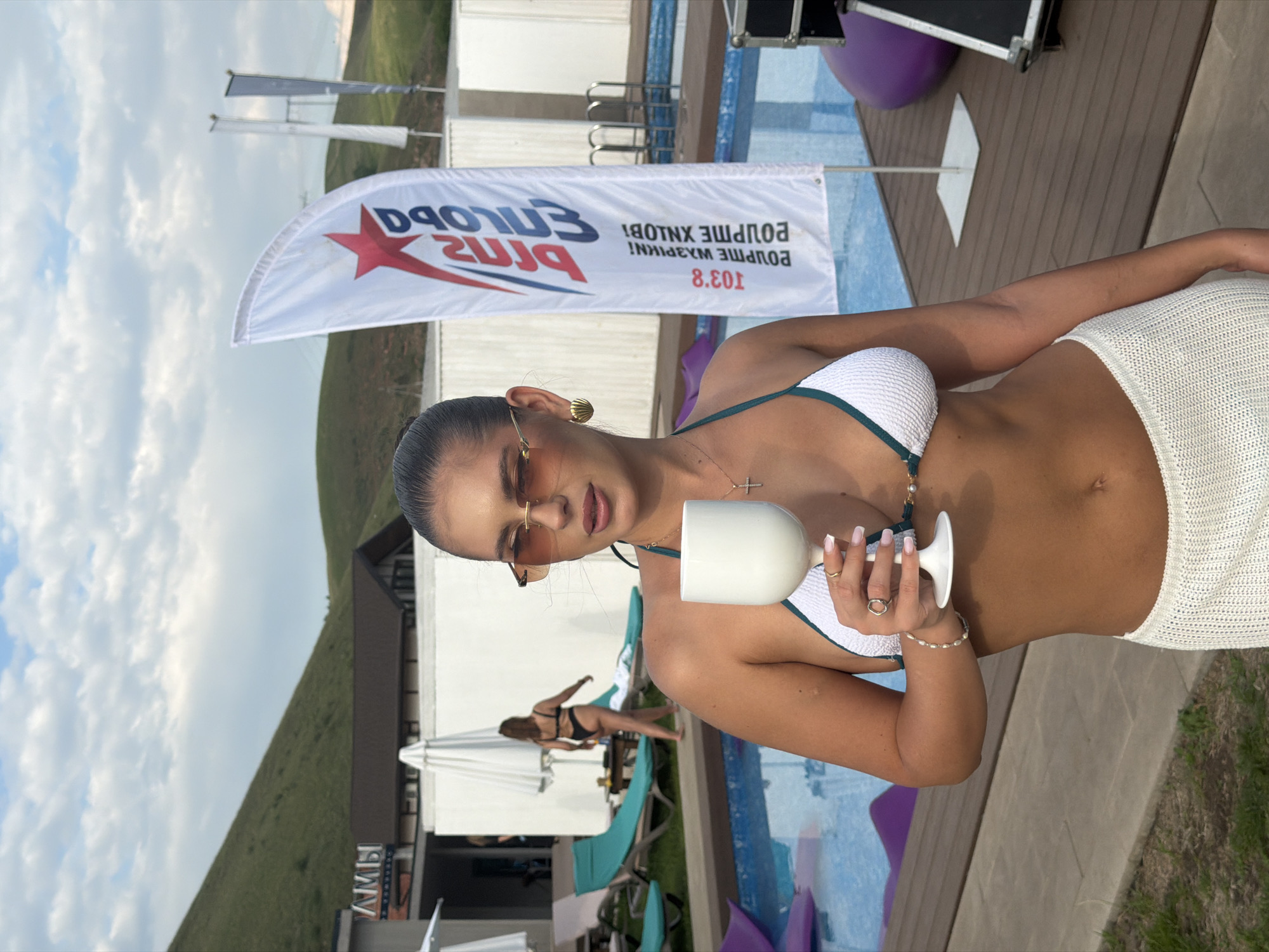 A woman wearing a white bikini top with green trim and a white knit skirt stands holding a white wine glass in front of a swimming pool. She has sunglasses on and is wearing a cross necklace and several rings. Behind her, a woman in a black bikini walks away from the camera. In the background, there are lounge chairs, umbrellas, and a white flag with a red star and text in Cyrillic. The scene appears to be at an outdoor venue, possibly a resort or club, near the city of Drokino, Russia, as indicated by the text on the flag. The sky is partly cloudy, and there are grassy hills in the distance.