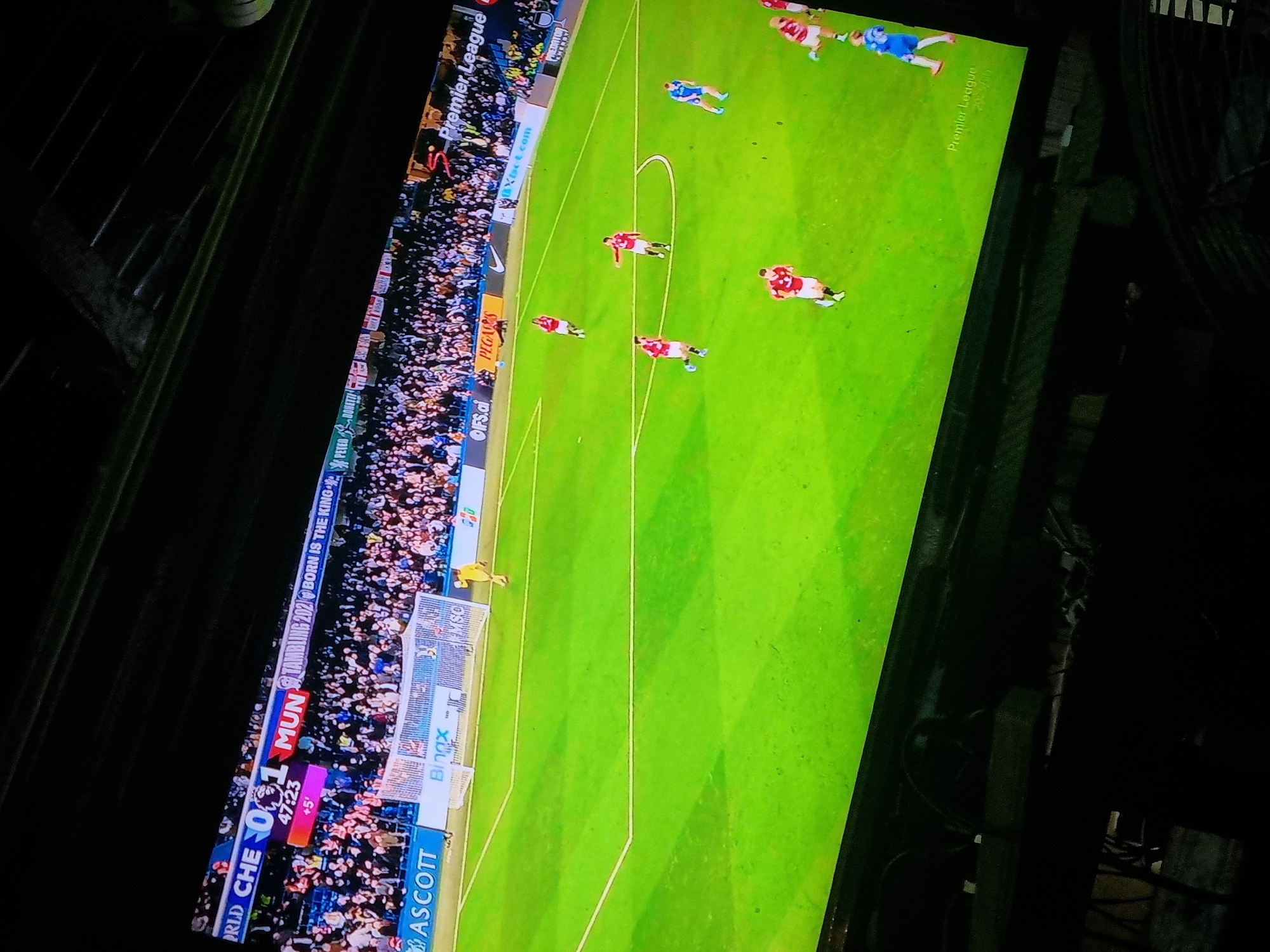 The image shows a television screen displaying a soccer game, specifically a Premier League match between Chelsea (CHE) and Manchester United (MUN). The score is CHE 0-1 MUN, and the game time is 47:23 with +5 minutes of added time. The screen also displays advertisements for "ASCOTT", "Bingx", "OIFS.CI PEGADES" and "Xscot.com". The Premier League logo is visible. The crowd is visible. There are some visible players and a goal keeper. The environment appears to be indoors with dark surroundings. There is no visible information to specify that the photo was taken in Onitsha, Nigeria.