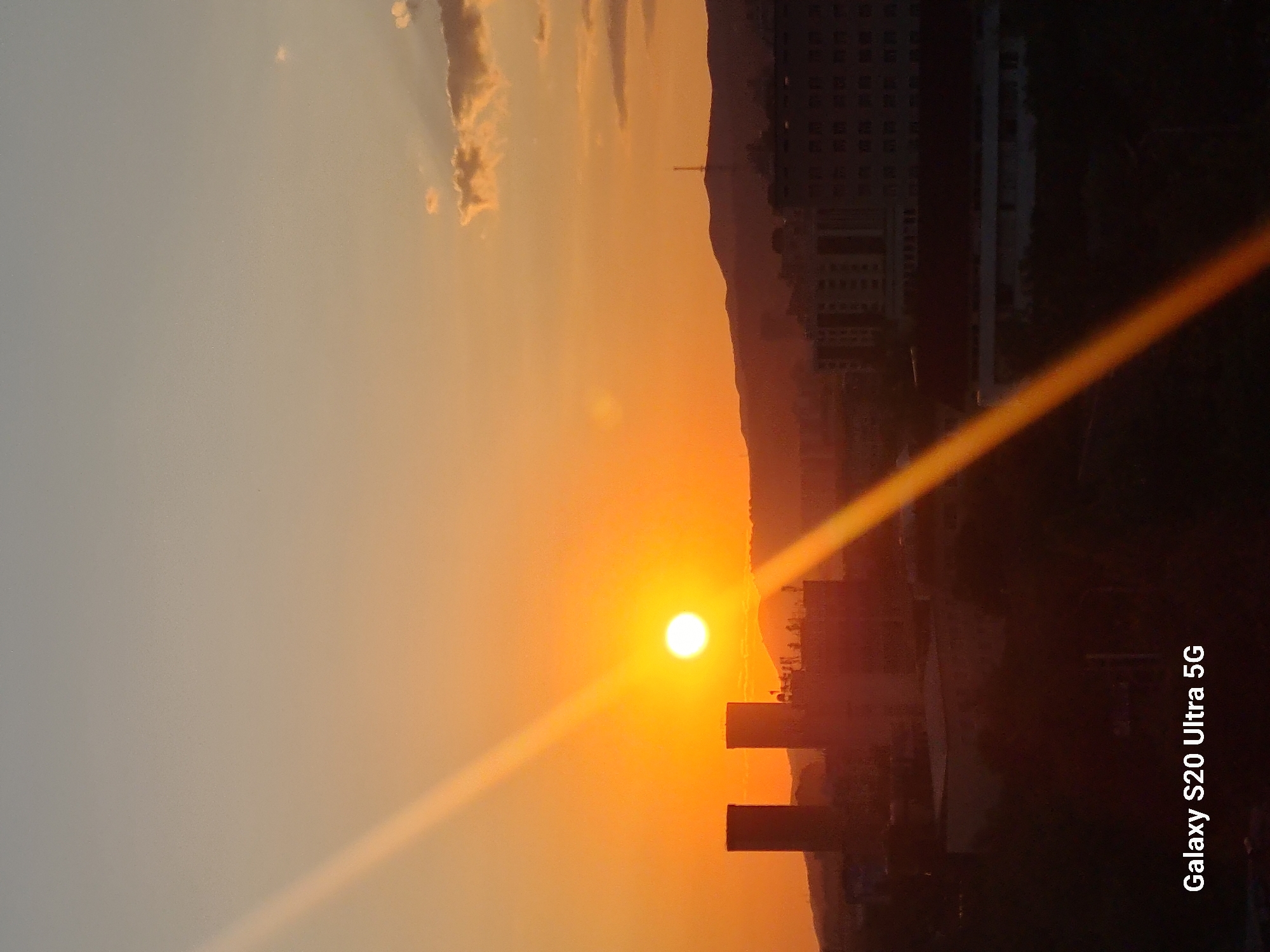 The image depicts a sunset scene over the city of Almaty, Kazakhstan. The sun, a bright orb, is positioned in the upper left quadrant, casting a warm orange glow across the sky. A prominent light flare streaks diagonally across the image from the upper left to the lower right. In the middle ground, silhouetted against the sunset, are several buildings and structures, including two tall, cylindrical towers and other multi-story buildings. The background features rolling hills and mountains, with a radio tower visible on one of the peaks. The foreground is dark and obscured, with the text "Galaxy S20 Ultra 5G" watermarked in the lower left corner. The overall atmosphere is one of twilight, with the city transitioning from day to night under the setting sun.
