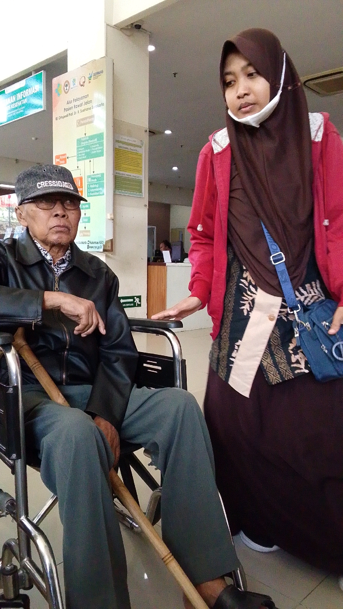 An elderly Indonesian man sits in a wheelchair indoors, possibly in the waiting area of a hospital or clinic in Pabelan, Indonesia. He wears glasses, a black jacket over a patterned shirt, and a dark cap. A walking cane rests against his right leg. Beside him, a younger Indonesian woman wearing a brown hijab and a red jacket stands, her right hand resting on the armrest of the wheelchair. She has a white mask pulled down below her chin. Behind them, a wall displays information posters in Indonesian, including one detailing patient service flow and another with text and graphics. The environment appears to be a well-lit, public indoor space with tiled flooring and fluorescent lighting visible on the ceiling.