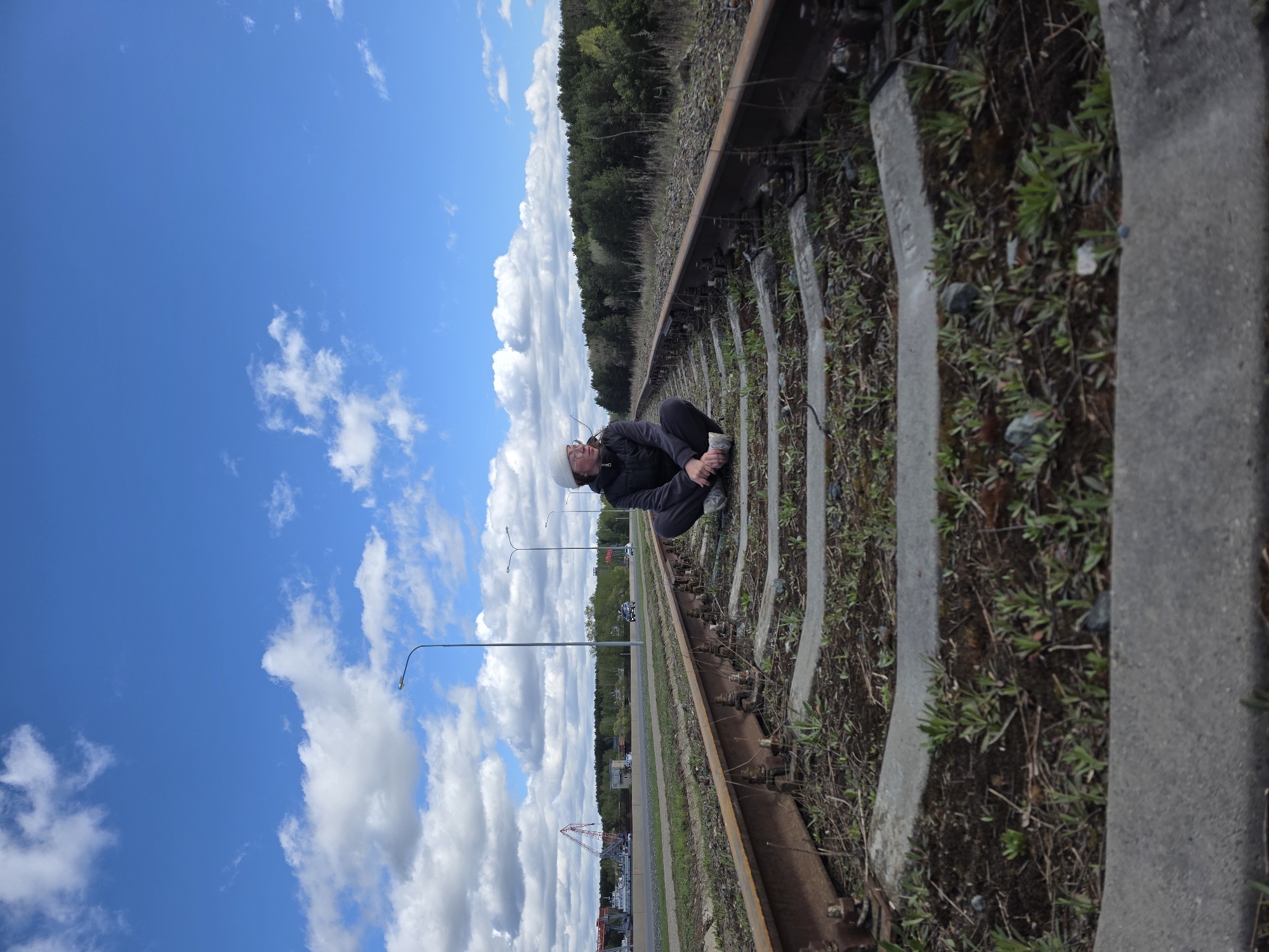 The image depicts a person squatting on a railway track under a blue sky with scattered clouds. The railway track runs parallel to a road, which is lined with streetlights and buildings in the distance. A forest is visible on the opposite side of the track. The location is in the city of Chaikovsky, Russia.