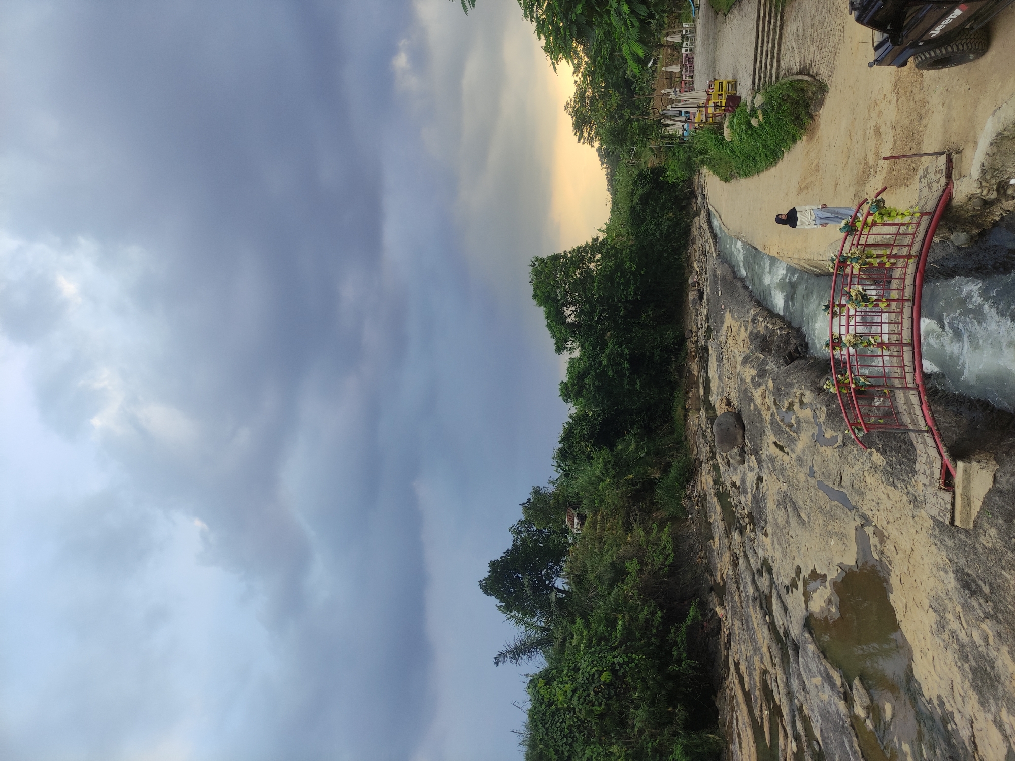 The photograph captures a landscape at Babakan Madang, Indonesia, under a predominantly cloudy sky. The ground slopes downwards, revealing a rocky terrain interspersed with water. Lush green vegetation lines the left side of the frame. A red-painted bridge with floral decorations is visible, with water flowing beneath it. A person is standing on the path near the bridge. A vehicle and some seating arrangements are located on the path higher up and to the right. The overall impression is that of a natural landscape with man-made structures integrated into it.