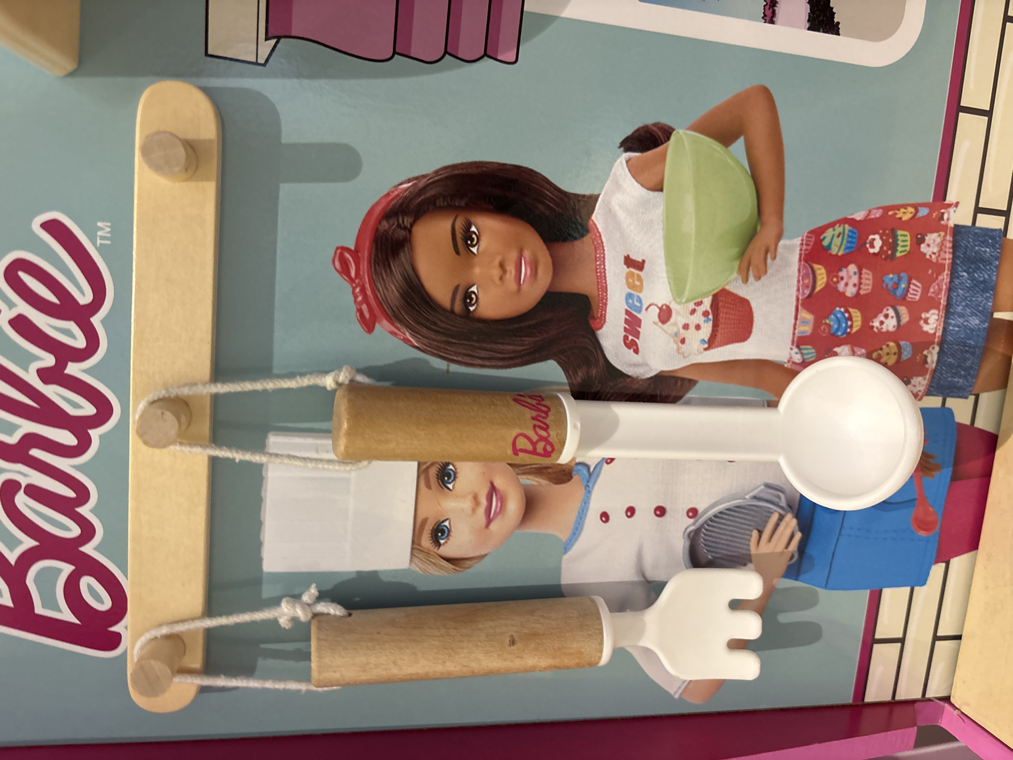 The image shows a close-up view of a Barbie toy kitchen set. Two Barbie dolls are depicted. The doll on the left is wearing a chef's hat and a white chef's coat with red buttons. She has blonde hair and blue eyes. The doll on the right has dark hair, brown eyes, and is wearing a red headband and a white t-shirt with the word "sweet" and cupcake graphics. She is holding a green bowl. Hanging from a wooden rack above the dolls are two kitchen utensils made of wood and white plastic: a fork-like utensil on the left and a large spoon on the right. The background features a light blue wall with graphics of cupcakes and kitchen items. The bottom of the image shows a wooden surface and a pink border. The image does not contain any information about the city of Dubai, United Arab Emirates.